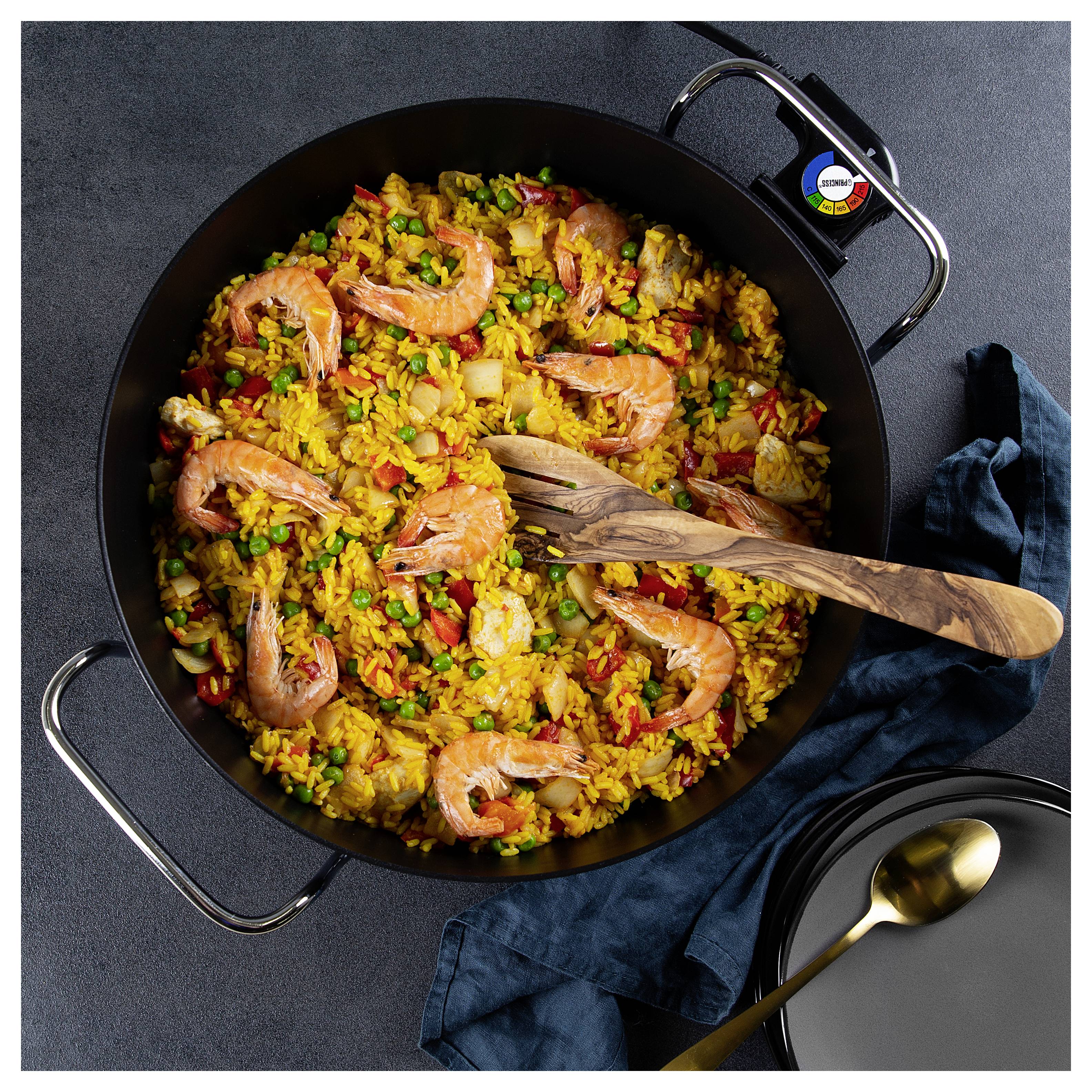 Överblick över en stor panna med räkpaella med ris, ärtor, paprikor och en träsked på ett mörkt bord.