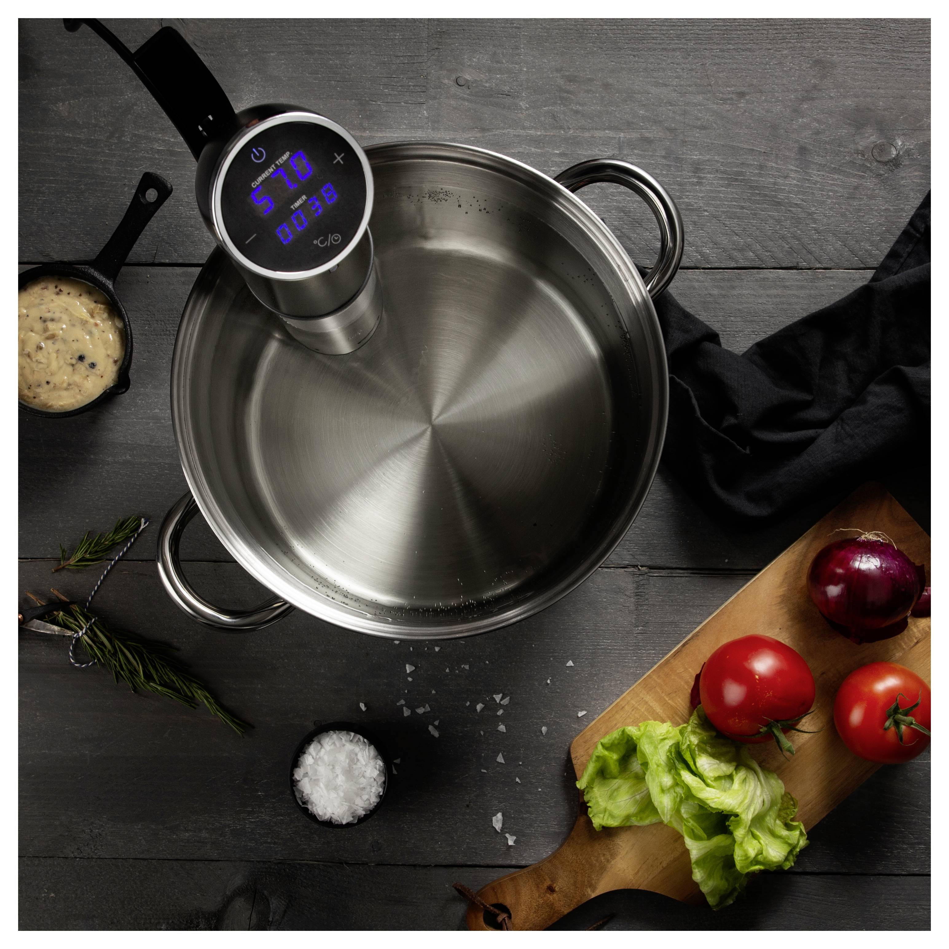 Sous vide-kokare med klämma fästad på en rostfri stålkruka på ett mörkt bord, med grönsaker och ingredienser arrangerade omkring den.