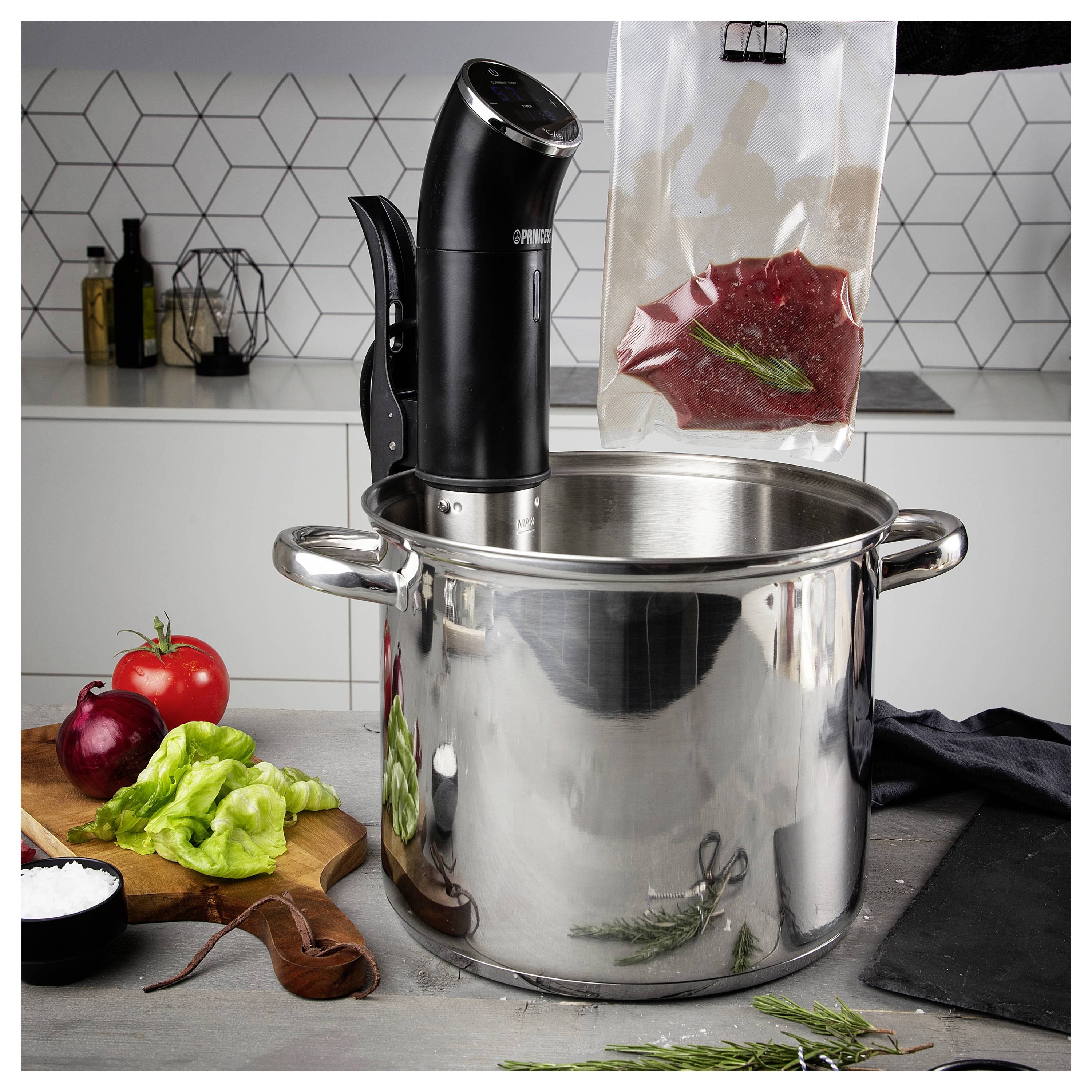 En nedsänkningscirkulatör värmer en stor rostfritt stålkruka för sous vide-matlagning, med en vakuumförseglad påse med kryddad kött som hänger bredvid den och ingredienser på bänken.