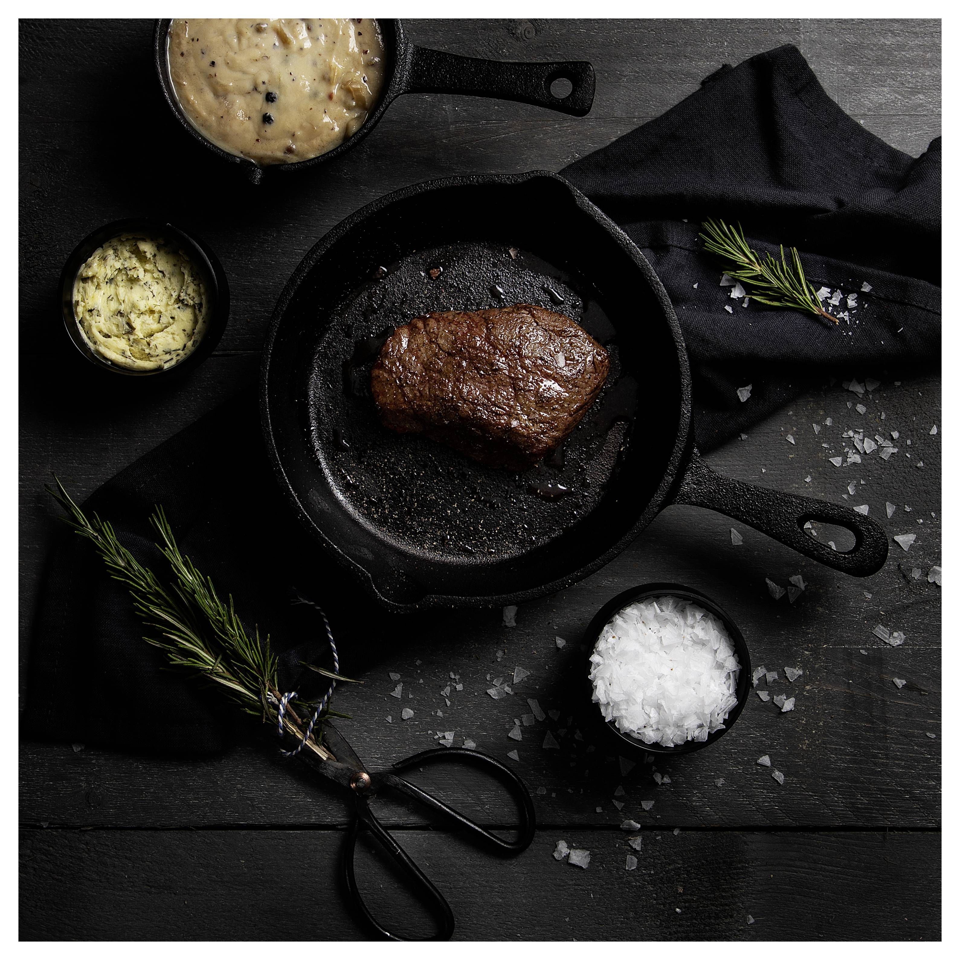 Stekt biff i en gjutjärnspanna på ett mörkt bord, omgiven av små skålar med sås, salt och rosmarinkvistar.