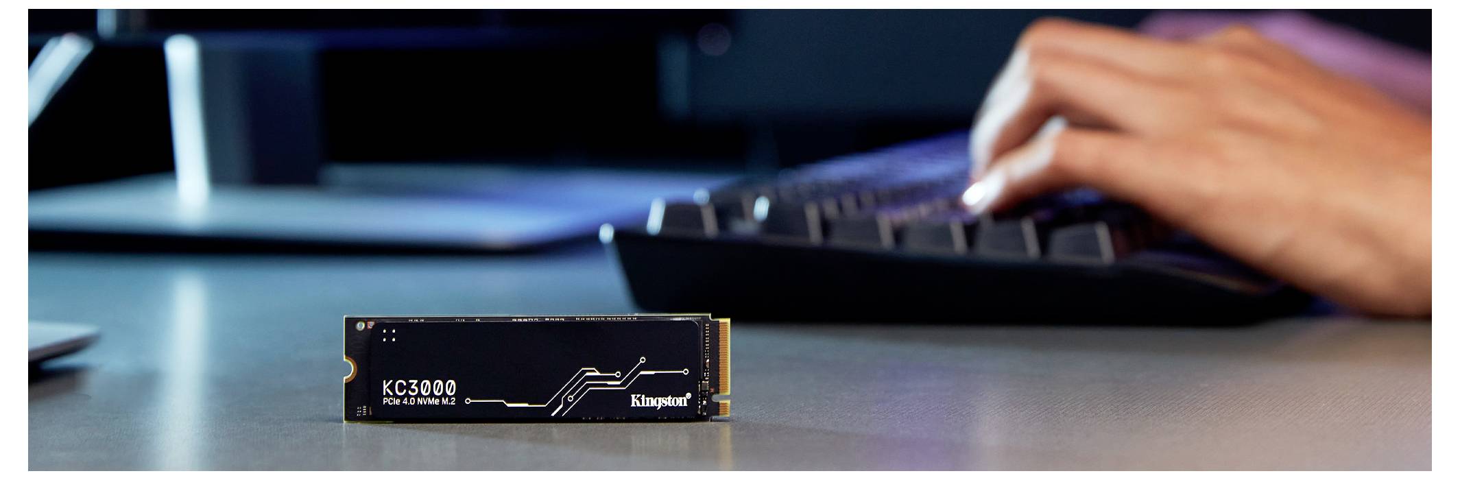 Närbild av en Kingston KC3000 SSD på ett skrivbord, med en person som skriver på en bärbar dator i bakgrunden.