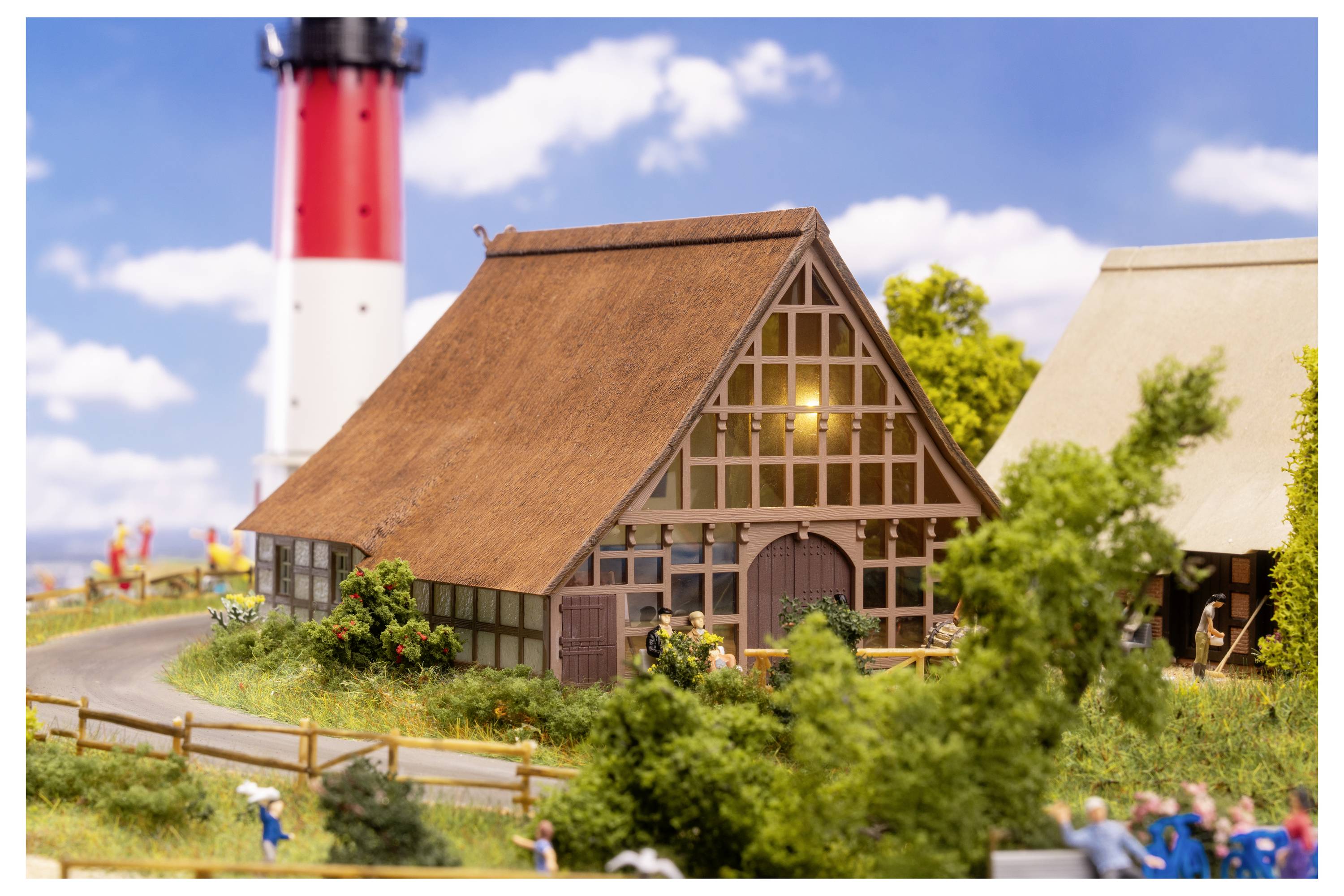 Miniatyrlandsbygd med ett strohtak hus vid en kurvande stig, röd-och-vit fyr i bakgrunden, och små figurer och träd i förgrunden.