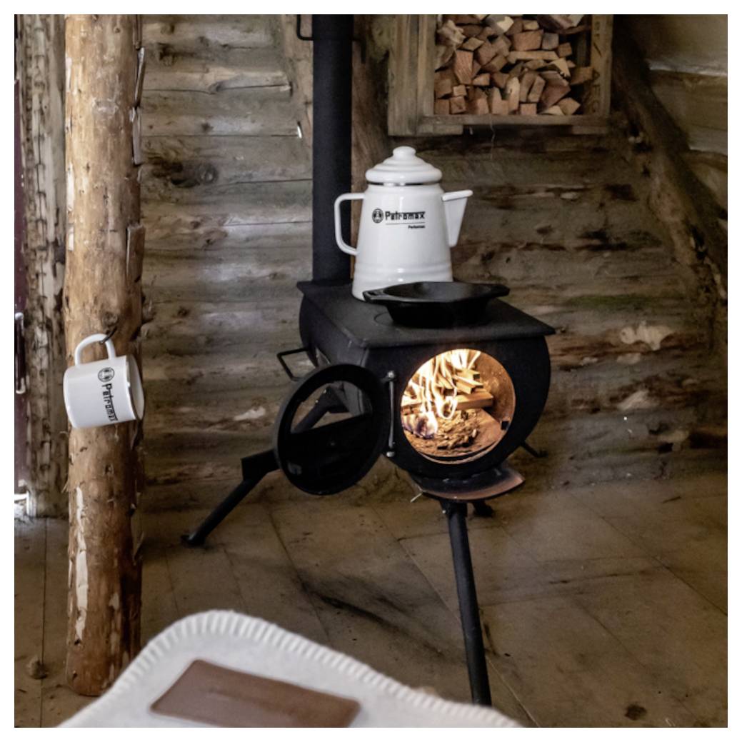Vedkamin värmer en rustik loggkoja, med en vattenkokare på toppen, en mugg på en stolpe och eldsvador glödande genom stövelns dörr.