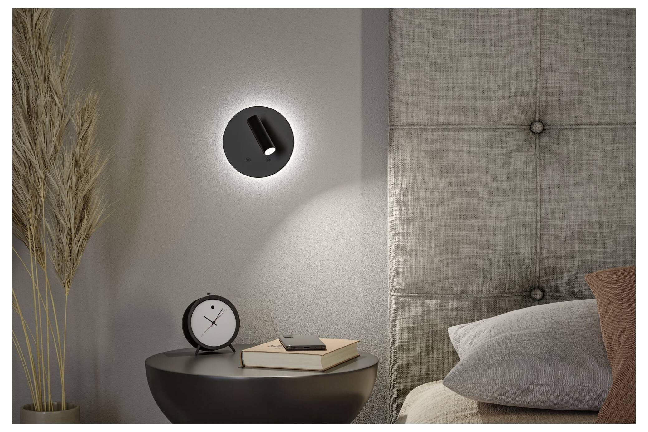 Modernt sovrumshjörn med en väggmonterad svart lampa ovanför ett litet runt sidobord som håller en klocka och böcker bredvid sängen och kuddar.