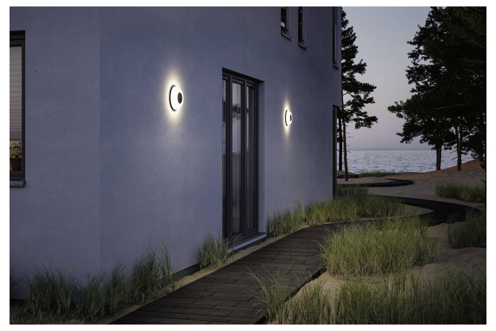 Modernt hus exteriör i skymningen med upplysta väggarmatur, en dörröppning, en träväg, gräs och utsikt mot vattnet och träden.