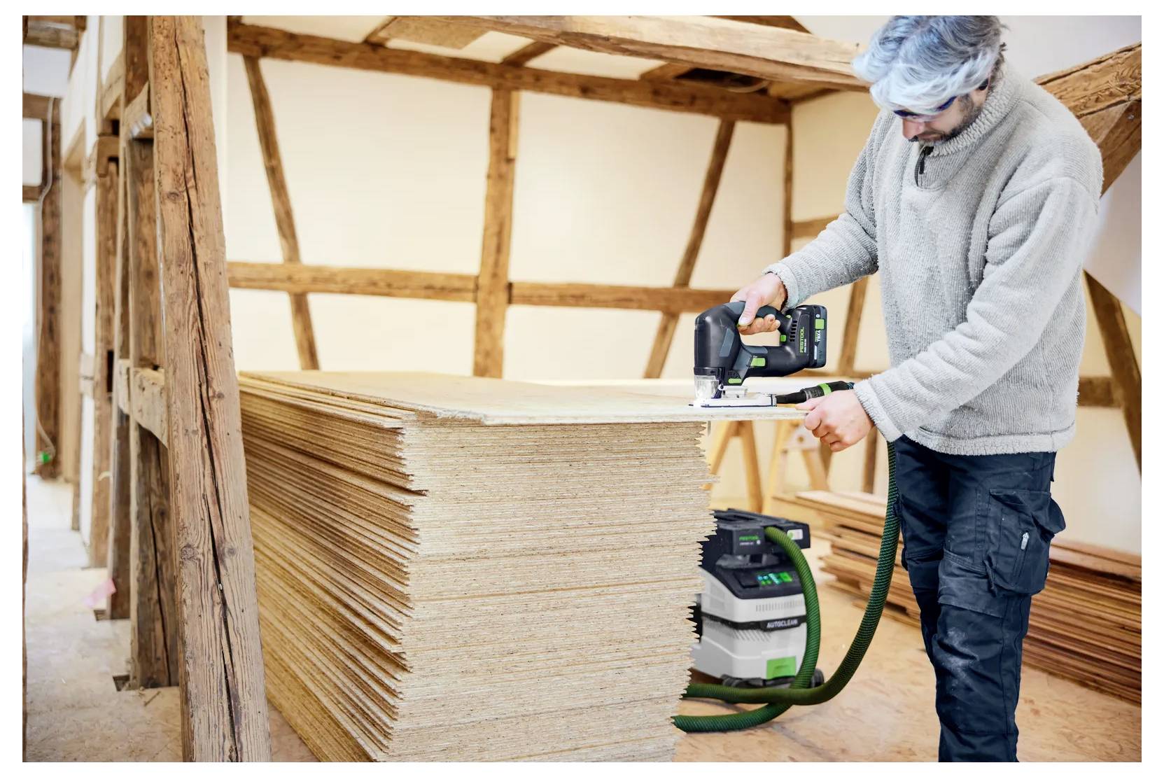 Arbetare som skär staplade plywoodskivor med en cirkulär såg i ett ofärdigt timmerrammat rum.