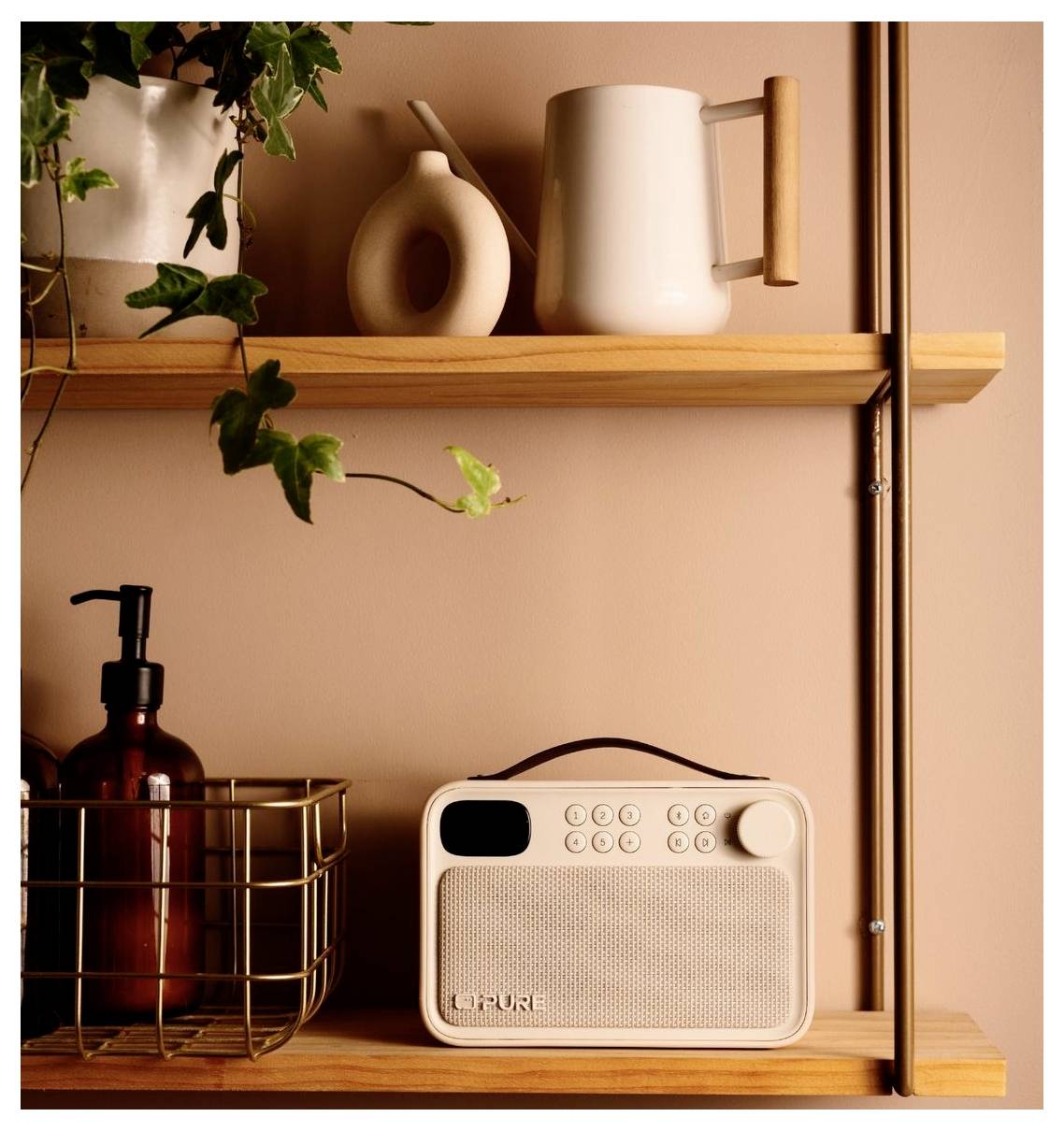 Liten beige bärbar radio på en trädhylla bredvid en tvålpump, med dekorativa vaser, mugg och hänga växt.