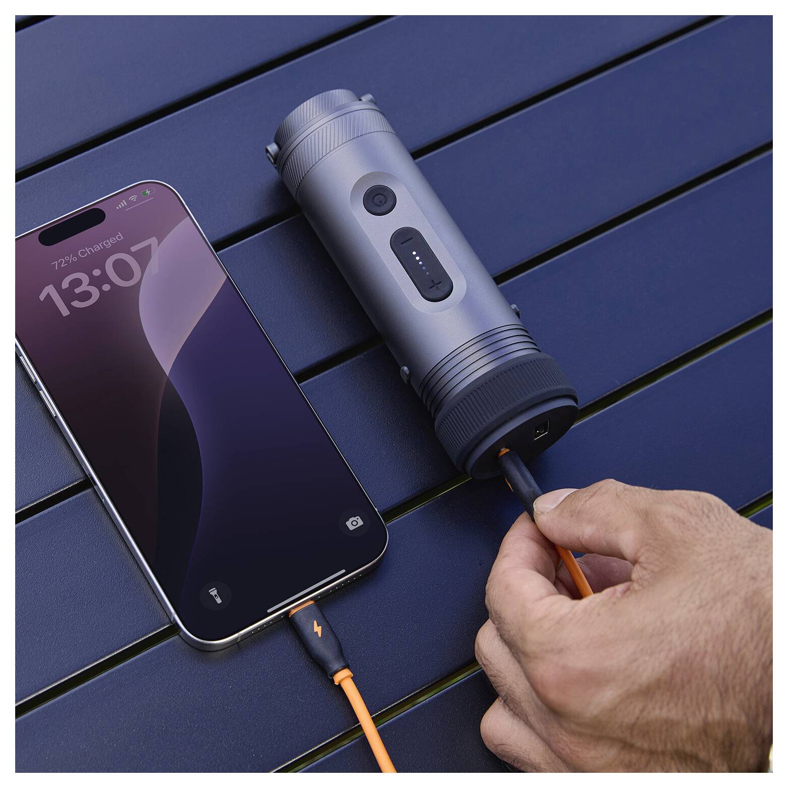 En hand sätter i en USB-C-kabel i en bärbar cylindrisk laddare bredvid en smartphone som laddar på ett mörkt bord.