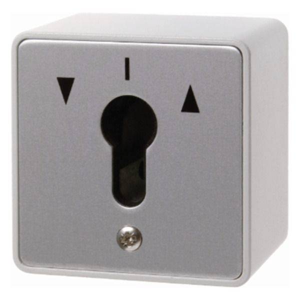 En kvadratisk silverfärgad nyckelväxlare med en nyckelformad öppning, upp- och nedpilpilar och en liten skruv nedan.
