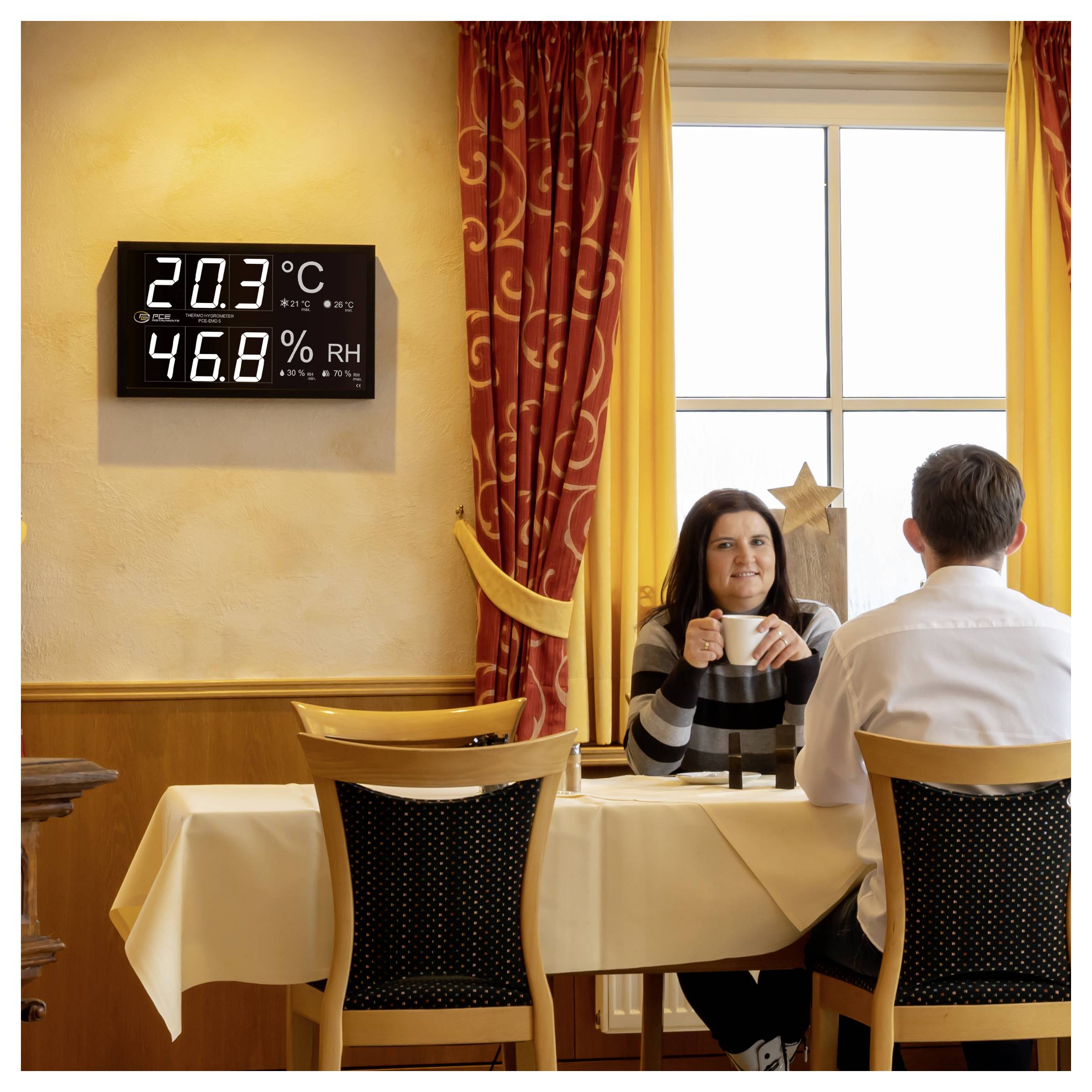 En kvinna och en man sitter vid ett bord på en restaurang nära ett fönster, medan en väggdisplay visar 20,3°C och 46,8% RH.