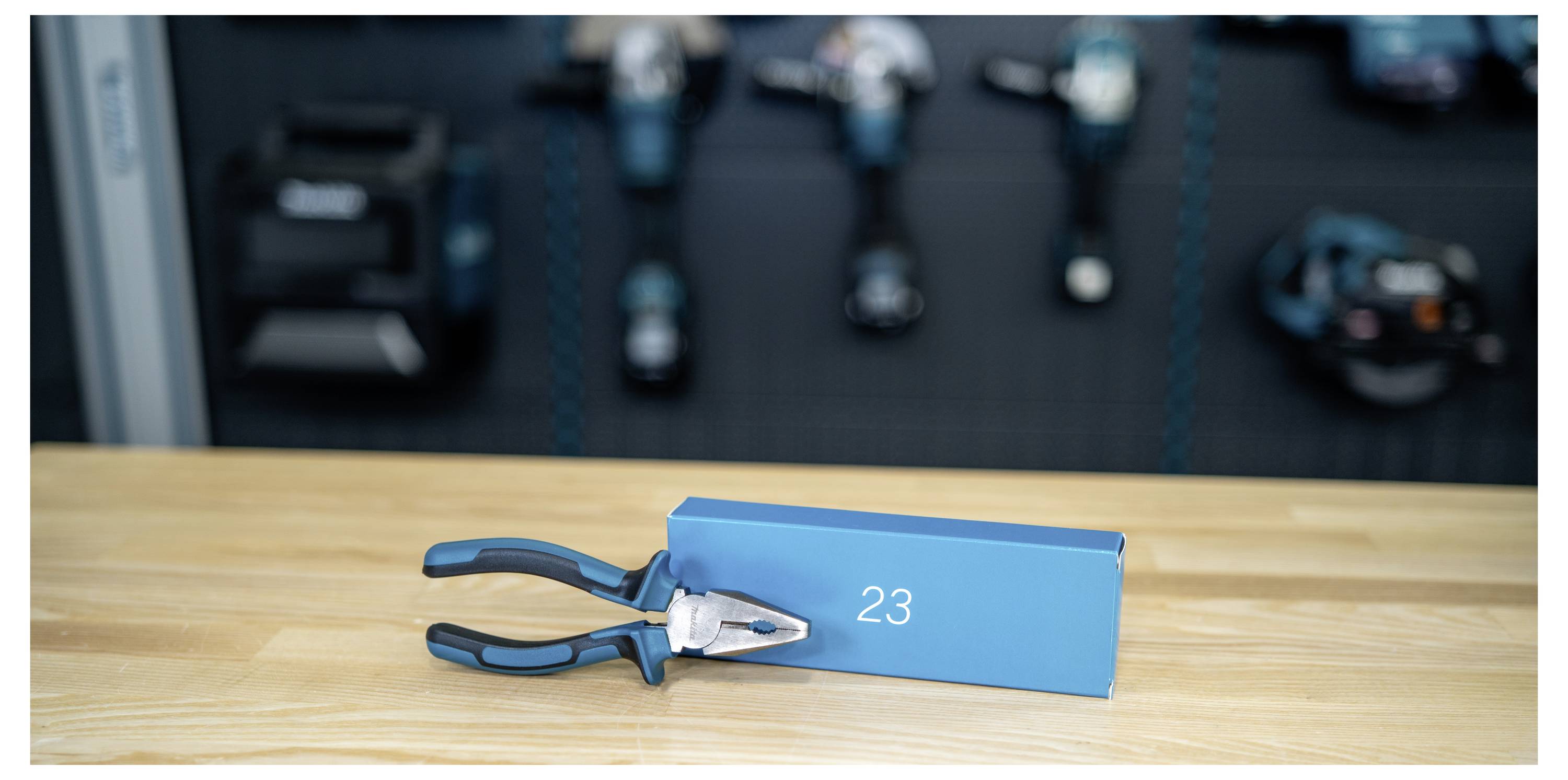 Blåhandtag tänger vilande på ett blått rektangulärt block märkt "23" på en träarbetsbänk, med suddiga verkstadsverktyg i bakgrunden.