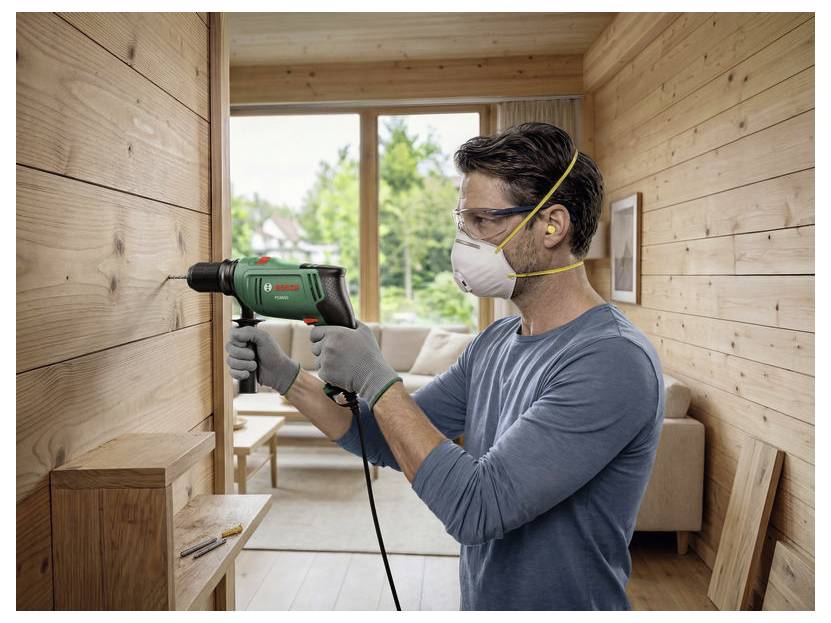 Man använder en eldrillborr för att installera träpanel inne i ett litet rum, klädd i handskar, skyddsglasögon och ansiktsmask.
