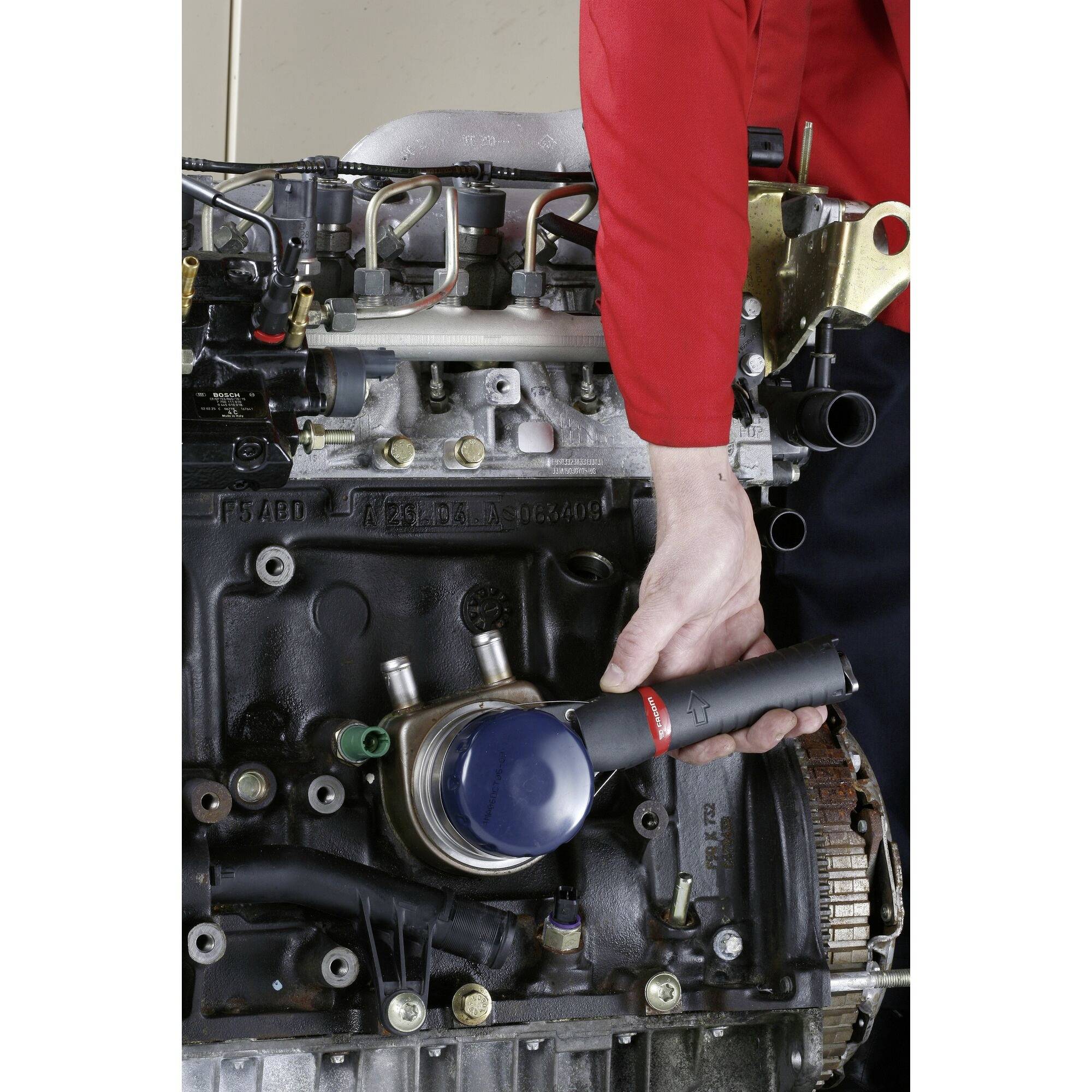 Mekaniker som håller ett cylindriskt verktyg nära ett motorblock, arbetar på framsidan av en exponerad bilmotor.