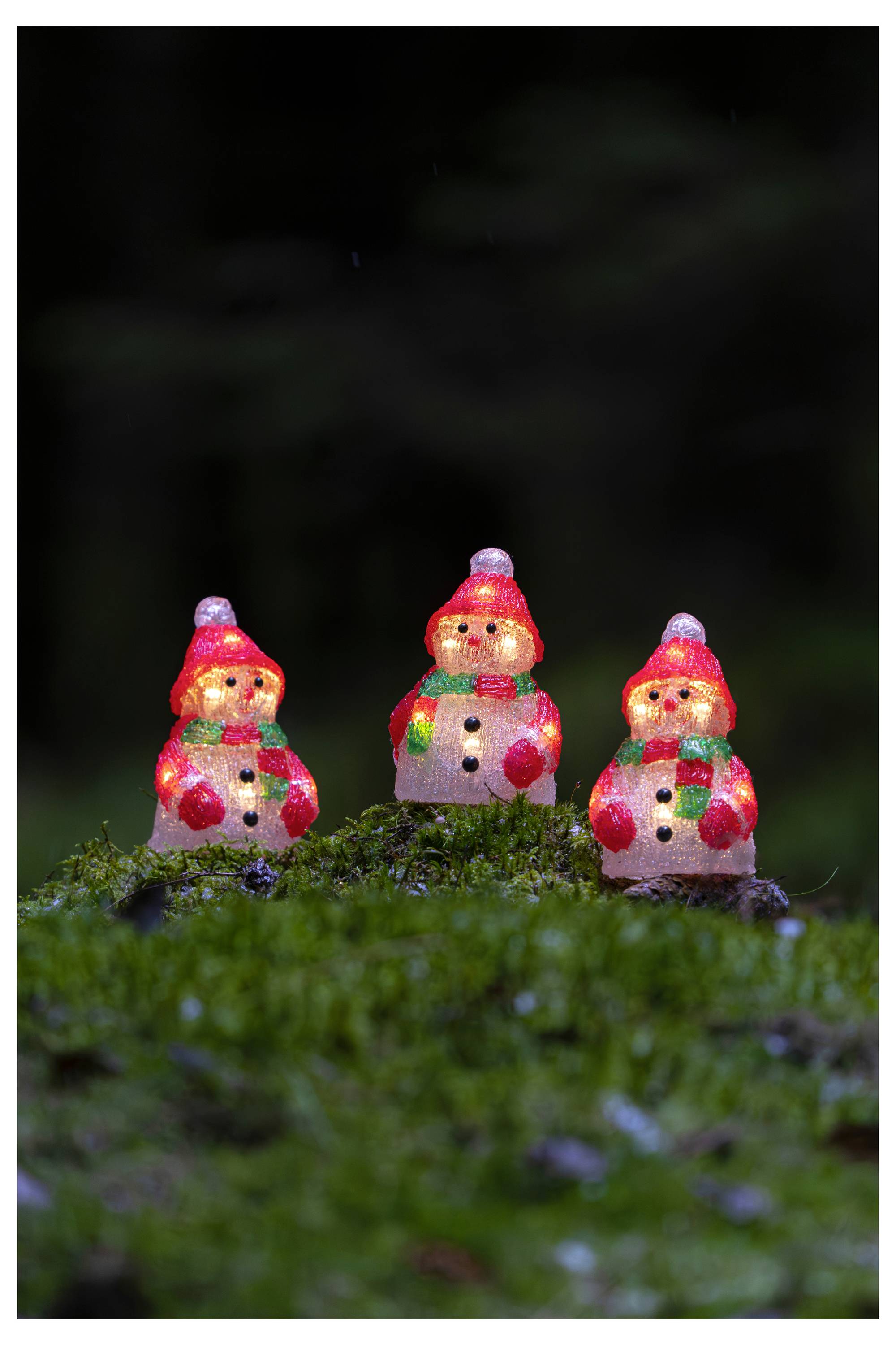 Tre små upplysta snögubbar sitter på mossa mot en mörk bakgrund.