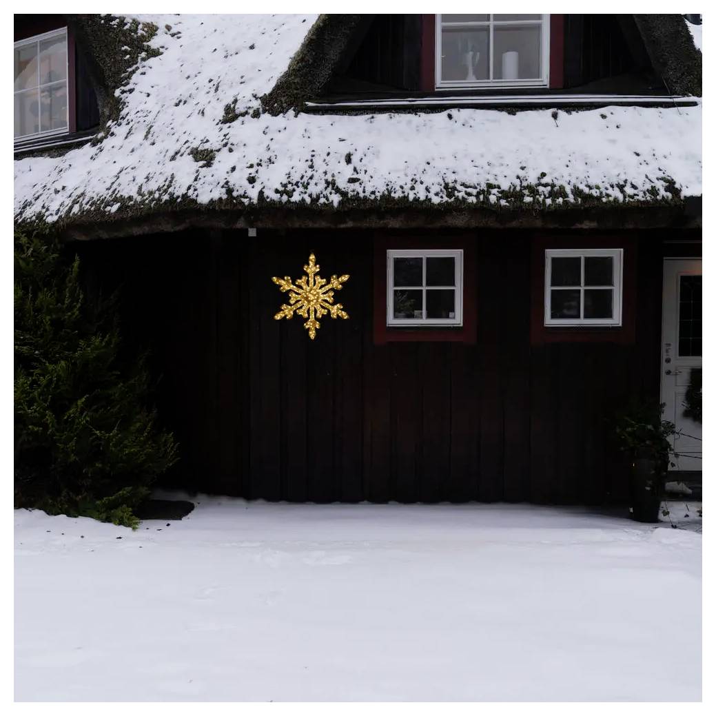 Snötäckt husfasad med en upplyst snöflakdekoraton på väggen, två fönster och en snöig trädgård framför huset.