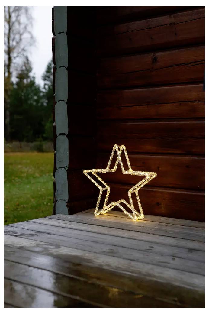 Glödande stjärnformad ljusdekoration på en träveranda bredvid en stockvägg, med en gräsbevuxen utomhusbakgrund.
