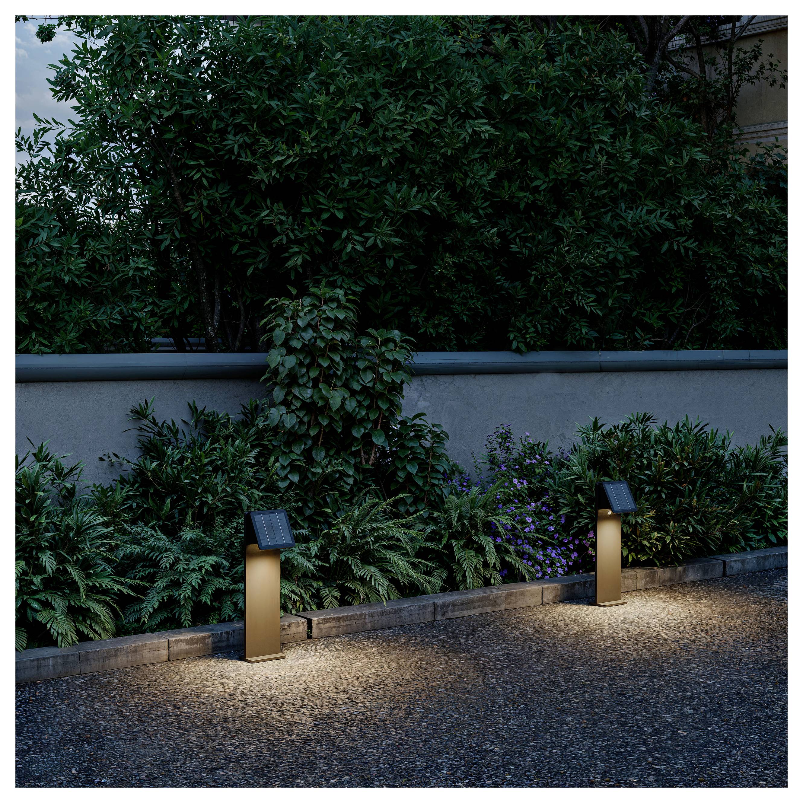 Två upplysta väg­stolpar belyser en stenlagd trädgårdsväg framför täta buskar och en vägg.