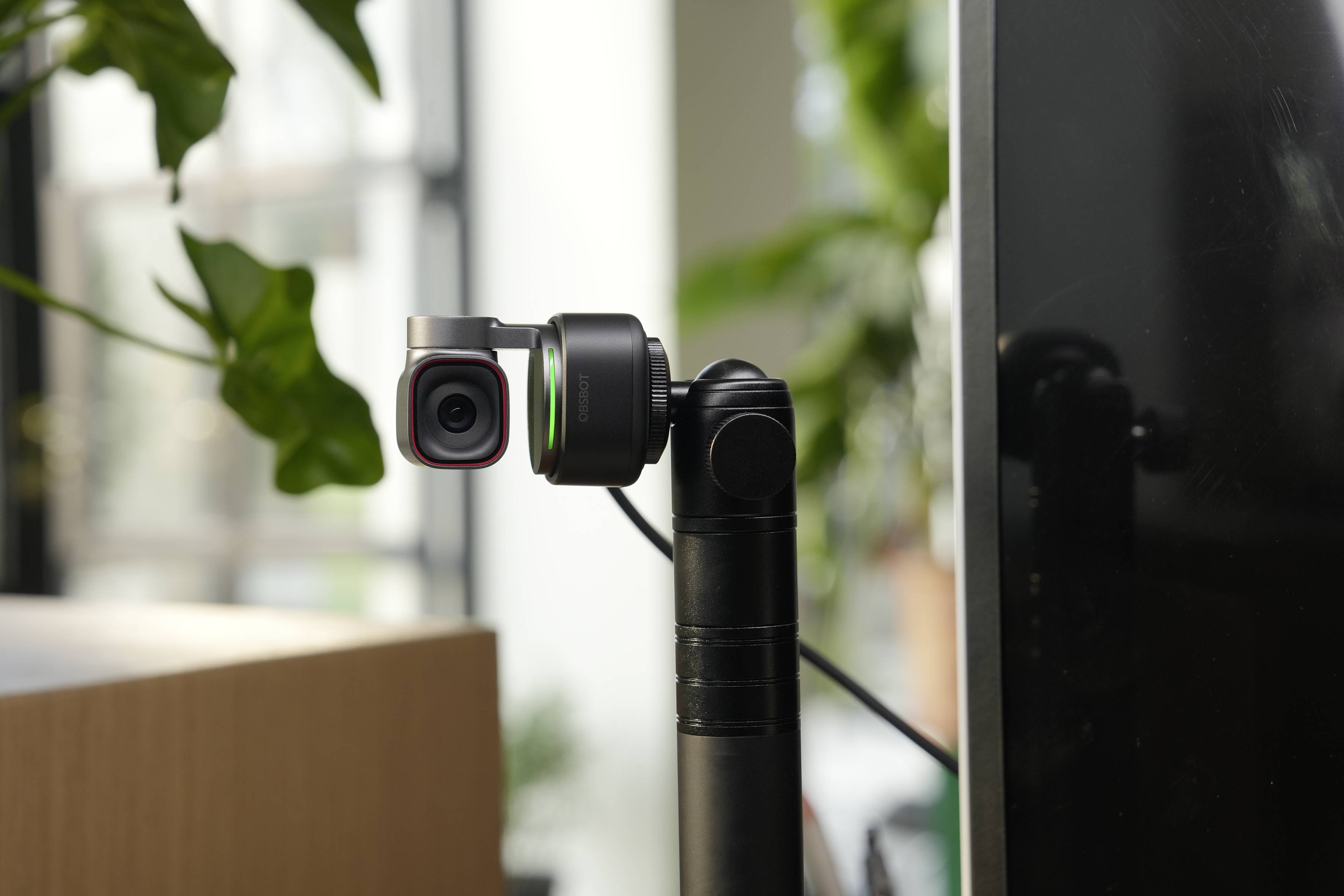 Liten actionkamera monterad på ett svart gimbal bredvid en monitor i en inomhus arbetsyta, med suddiga växter i bakgrunden.