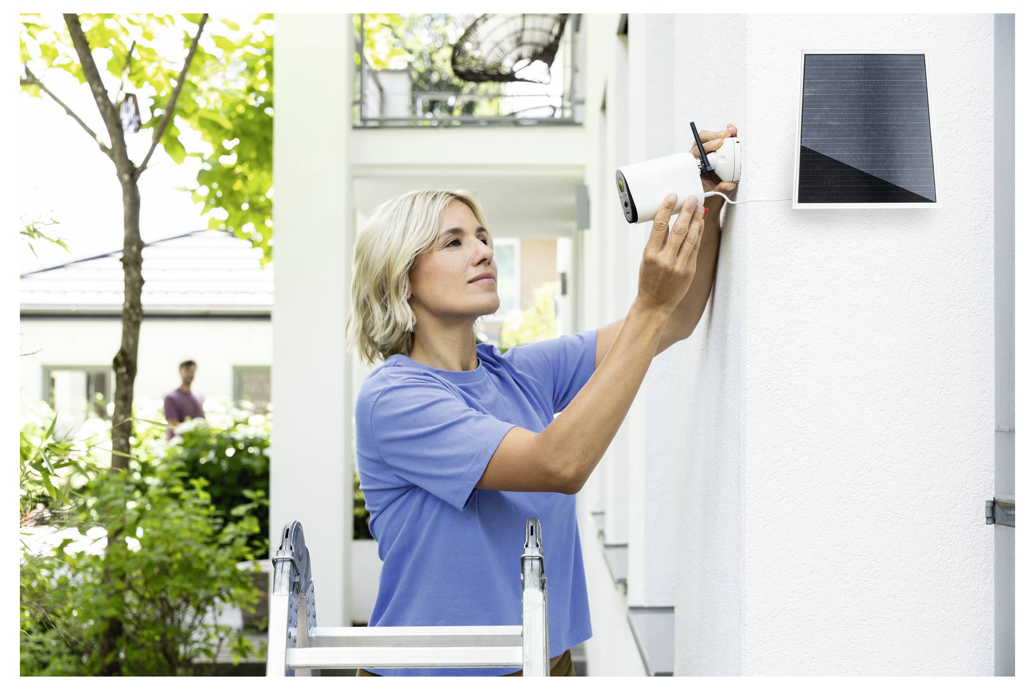 Kvinna på en stege som installerar ett utomhus säkerhetskamera bredvid en väggmonterad solpanel utanför ett hus.