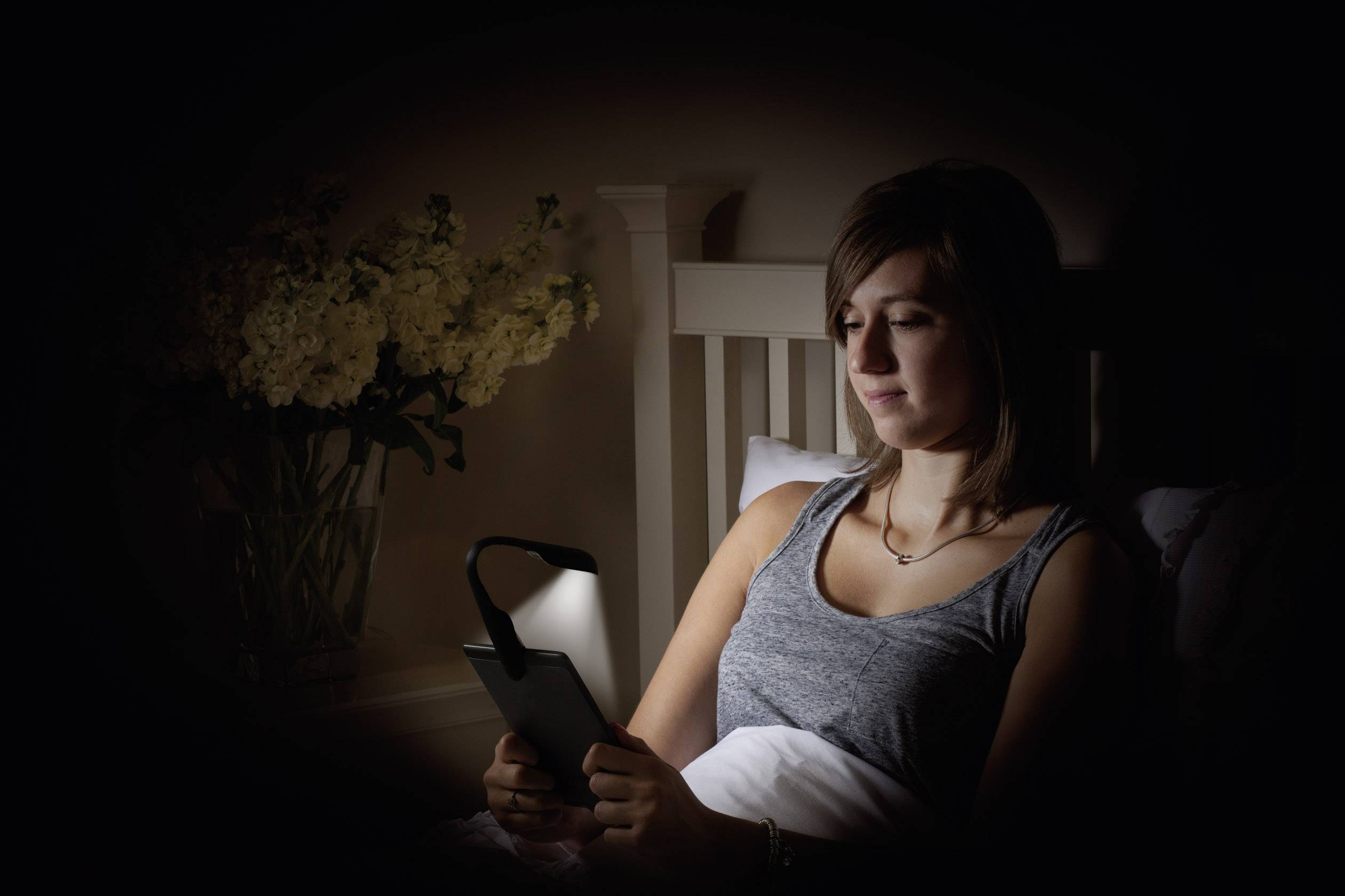 En person läser i sängen på en e-läsare medan en lampa lyser upp skärmen. I bakgrunden står en vas med blommor.