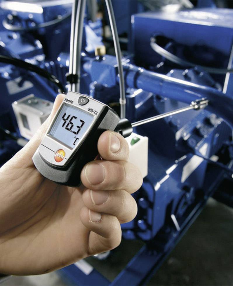 En hand håller ett digitalt termometer framför en blå maskinanläggning, där termometern visar en temperatur på 46,3 °C.
