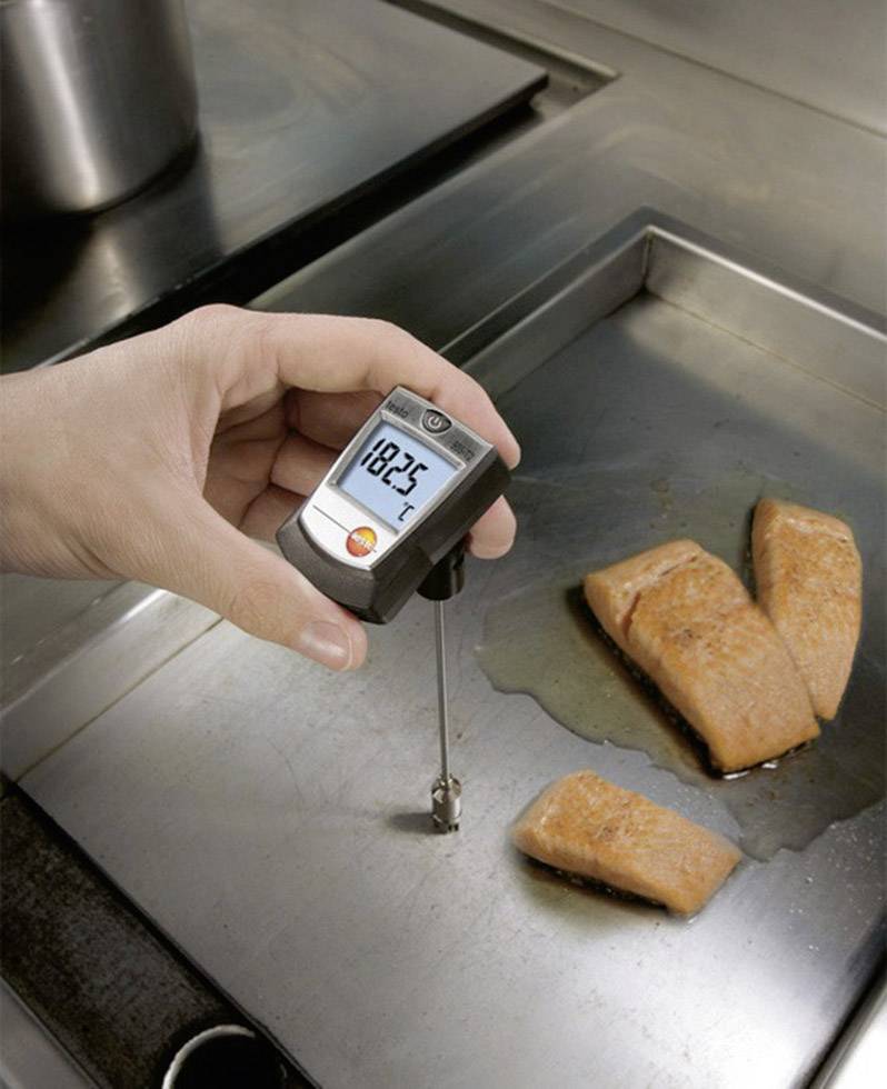 En hand håller ett köttermometer som visar en temperatur på 182,5 grader intill tre laxfiléer som ligger på en het stekpanna.