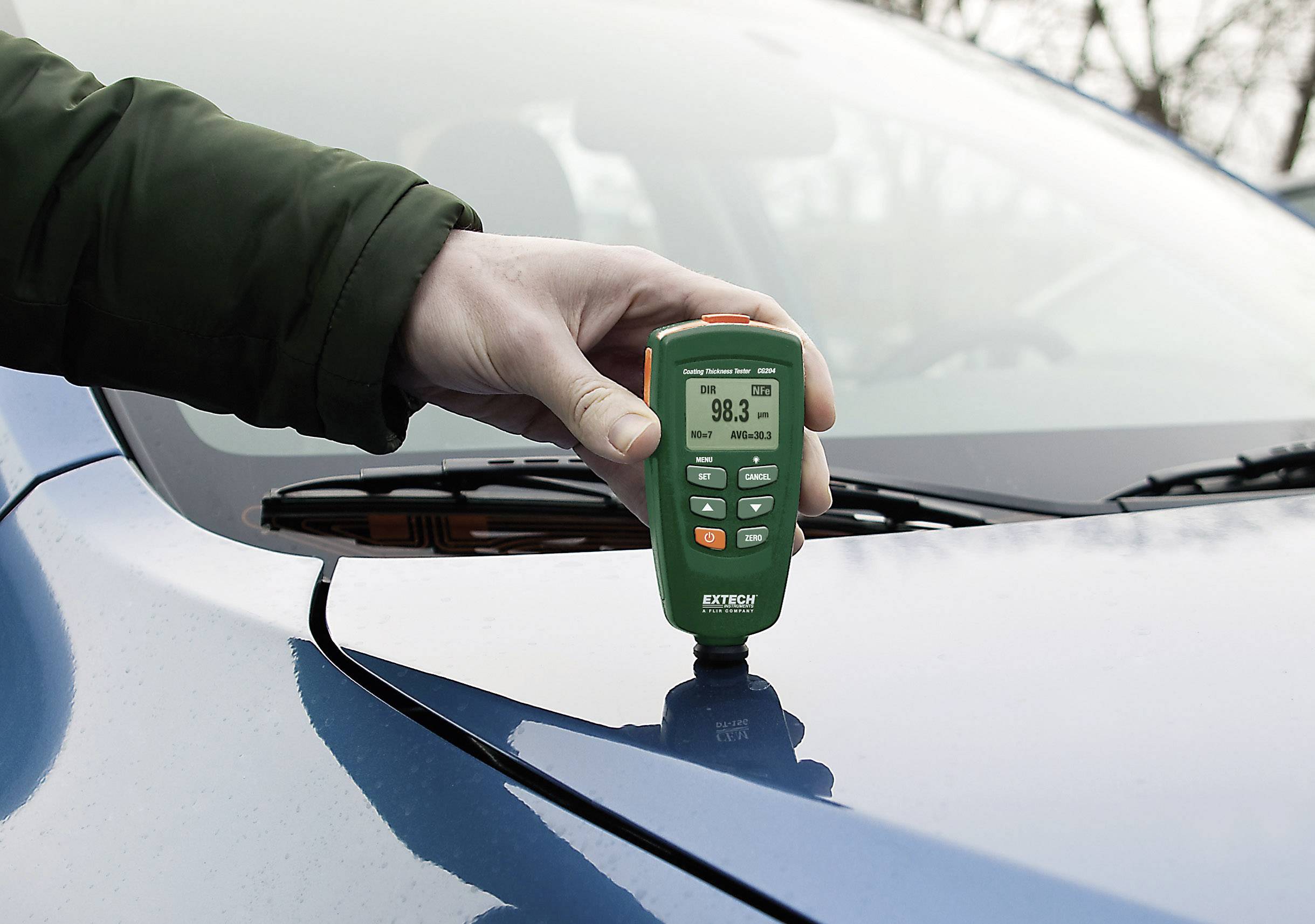 En hand håller ett tjockleksmätningsinstrument mot ett blått biltak som visar en tjocklek på 98,3 mikrometer.