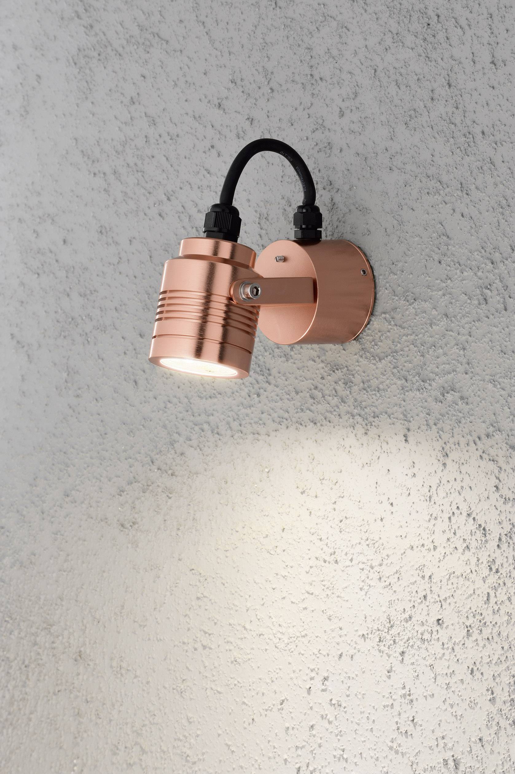 Kopparfärgad vägglampa på en strukturerad, ljusgrå vägg som lyser nedåt och belyser ytan.