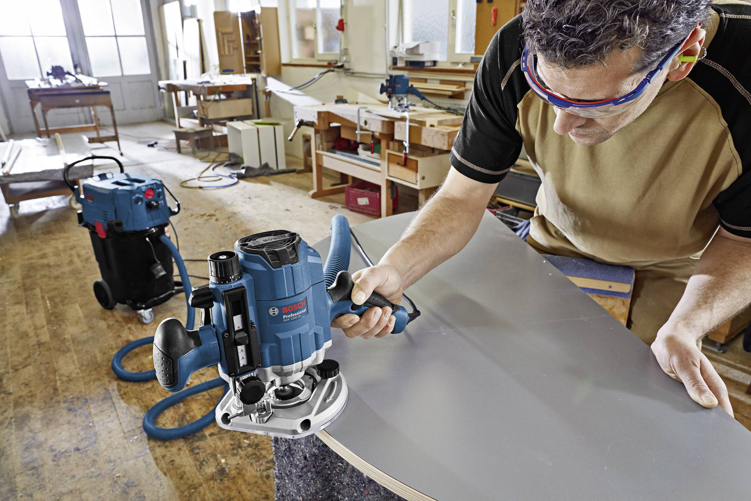 En hantverkare bearbetar en stor träskiva med en eldriven maskin i en verkstad. I bakgrunden syns ytterligare verktyg och arbetsbänkar.