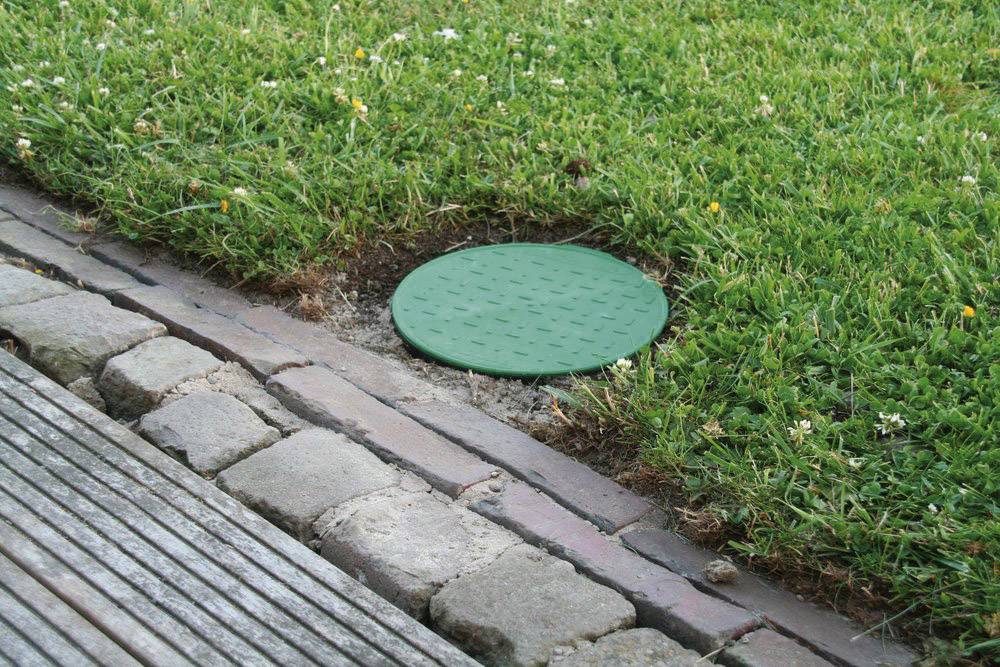 En grön kabelbrunn på en gräsmatta, inbäddad mellan trottoarstenar och en betongväg, omgiven av gräs och små blommor.