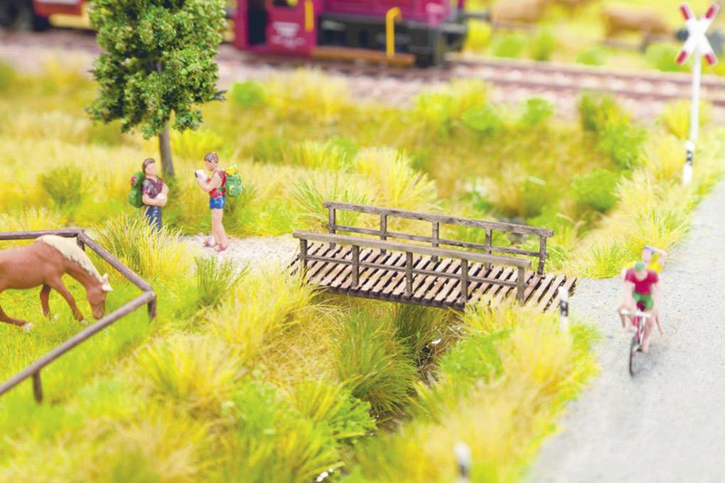 En miniatyrscen visar två vandrare nära ett träd, en häst på ängen och en person på en cykel bredvid en liten träbro. I bakgrunden passerar ett tåg.