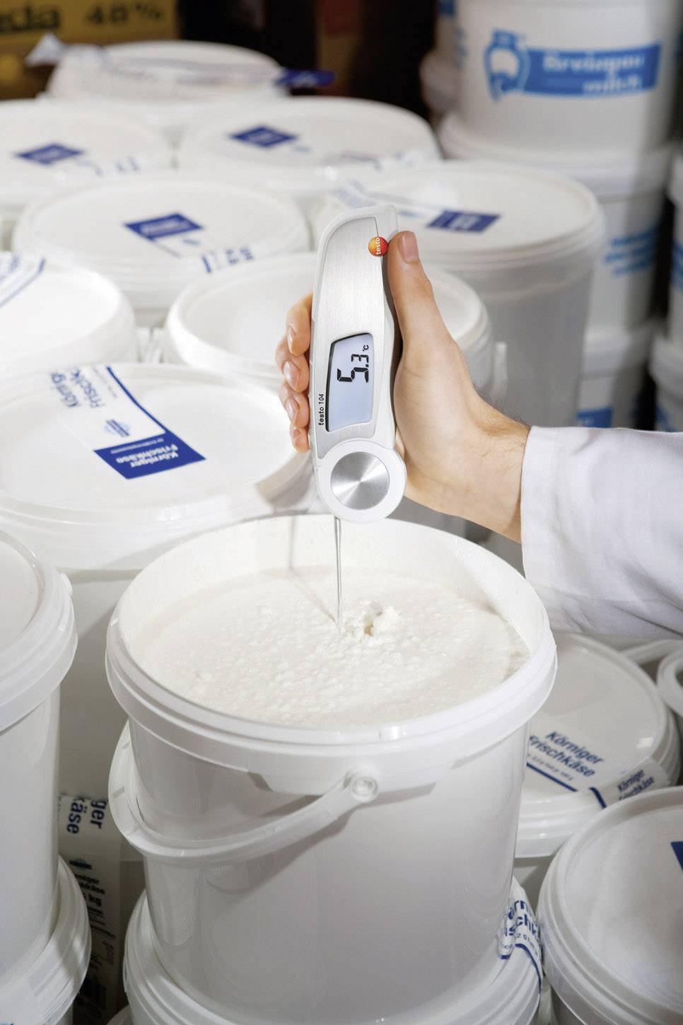 En person mäter temperaturen på en vit krämig substans i en öppen hink med en digital termometer.