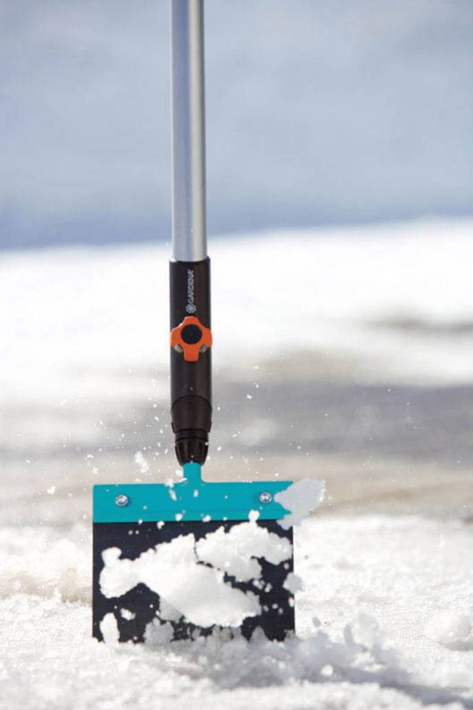 Snöskyffel med långt skaft lyfter snö från marken. Fokus på snöns rörelse. Vinterlandskap i bakgrunden.