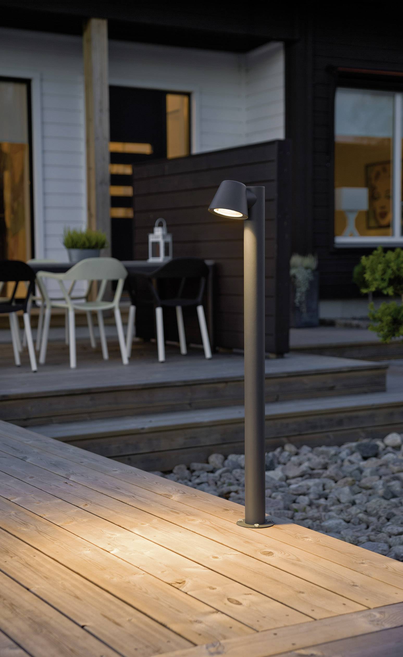 Svart-grå utebelysning lyser upp trätrall bredvid terrass med bord, stolar och lykta framför mörkt hus vid skymning.