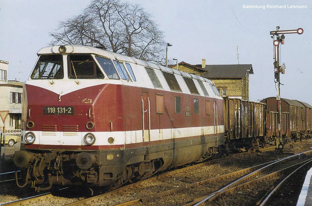 En röd diesellok drar ett godståg genom en järnvägsstation. I bakgrunden syns träd och en signal.