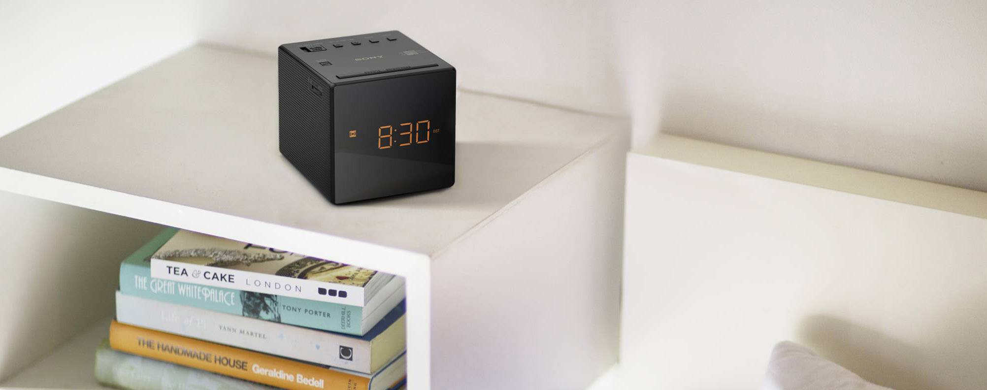 En svart digital väckarklocka visar '8:30' och står på en vit hylla bredvid en hög med böcker, däribland 'Tea & Cake'.