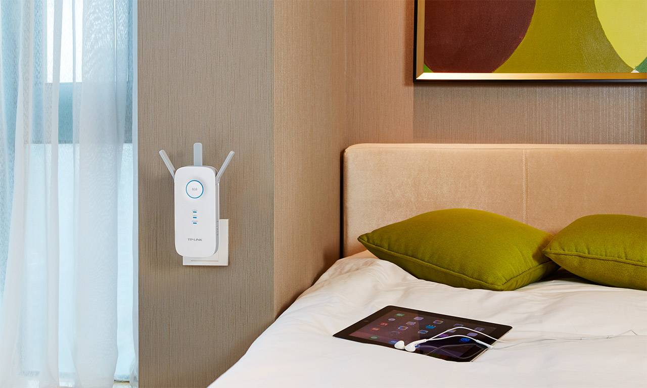 En WiFi-repeater är installerad på väggen i ett sovrum intill en säng med gröna kuddar. En surfplatta ligger på sängen.