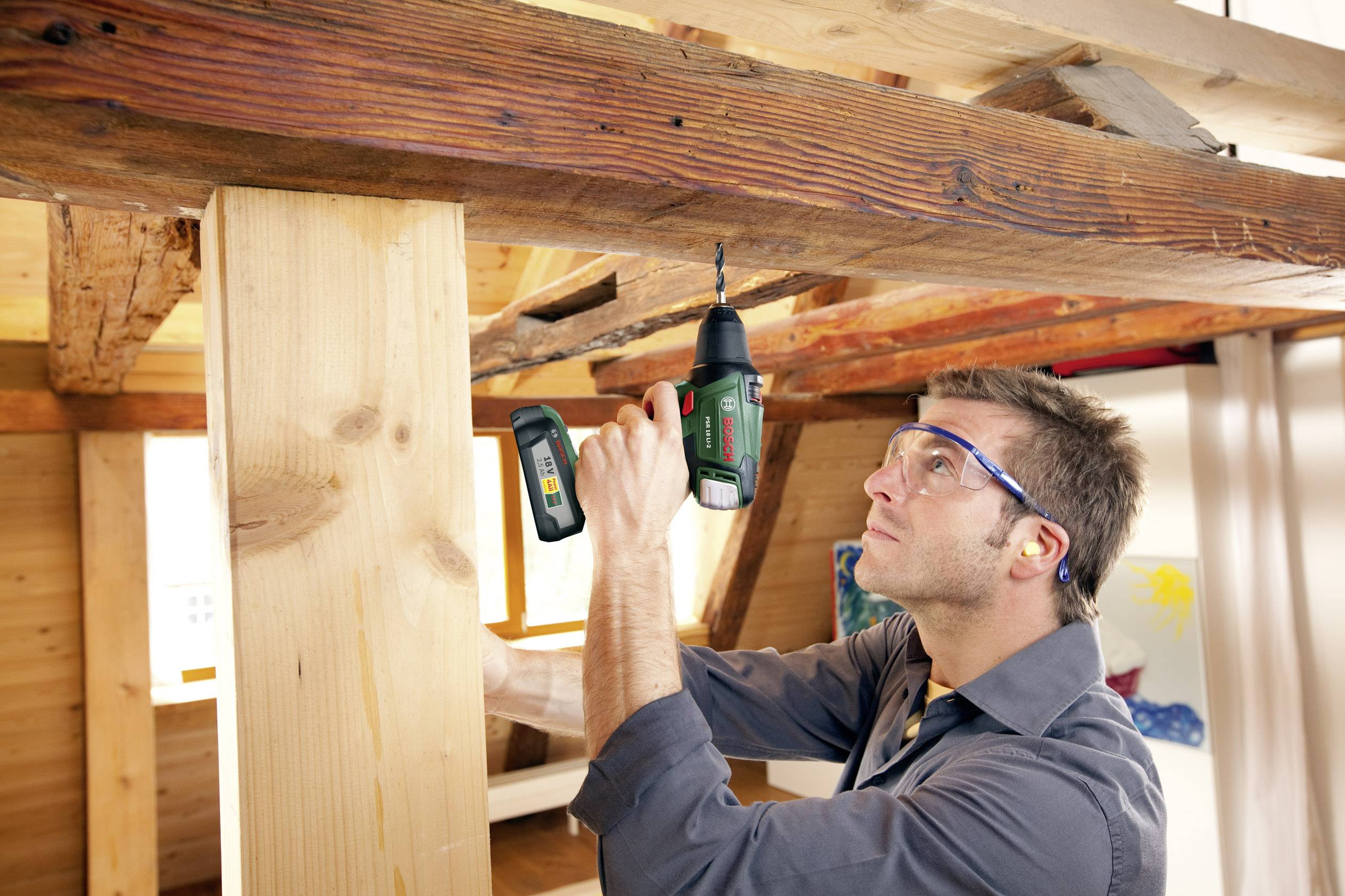 En man med skyddsglasögon och hörselskydd använder en borr för att borra ett hål i en balk i ett rum med träpanel.