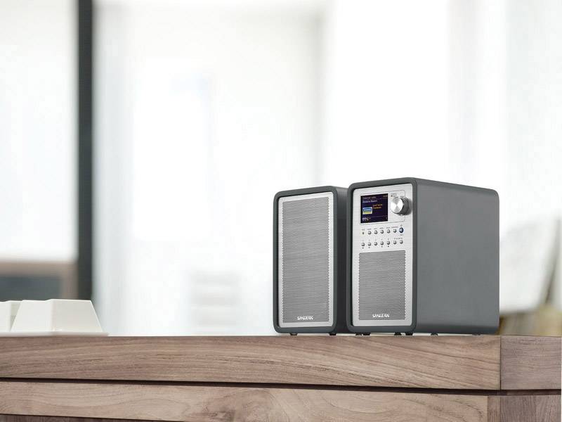 Två kompakta högtalare på ett träbord, modernt design, med en oskarp interiör i bakgrunden.