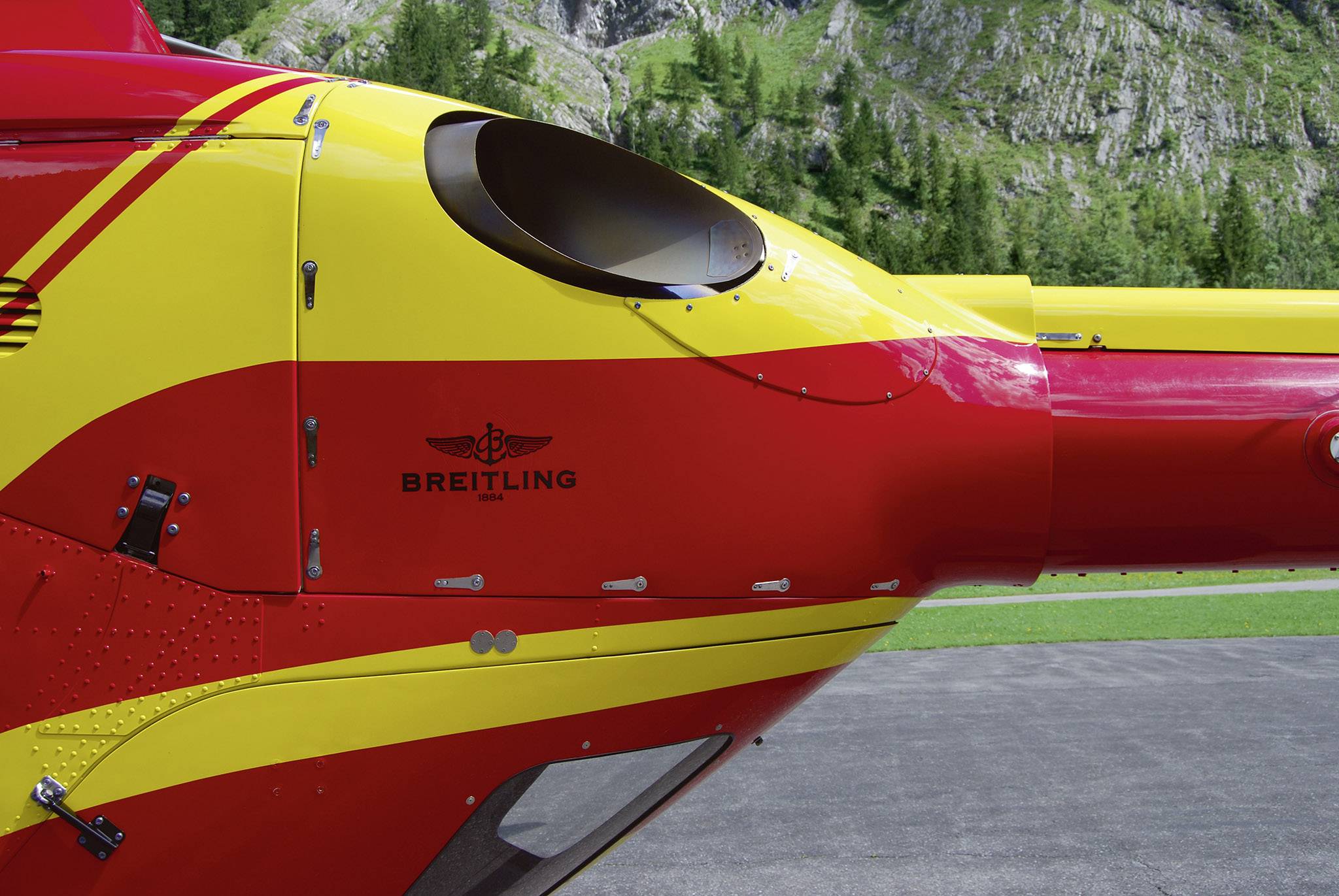 Närbild av en röd-gul helikopter med Breitling-logotyp på skrovet, parkerad på en asfalterad yta, med berg i bakgrunden.