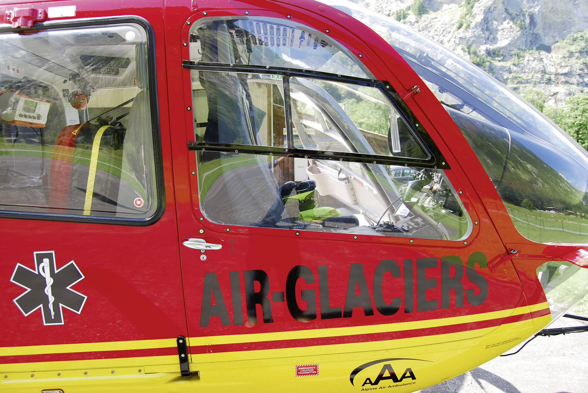 Röd helikopter från Air-Glaciers, med medicinskt emblem, står på en landningsplats. I bakgrunden syns klippor.
