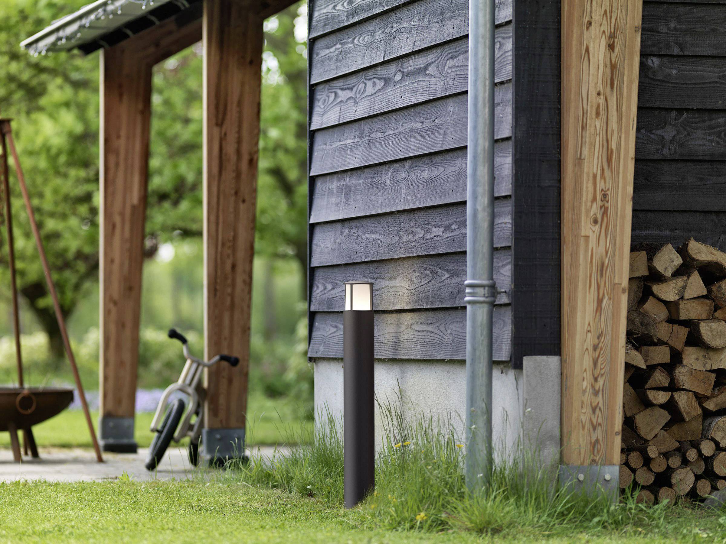 En liten svart stolpe med ljus bredvid ett vedskjul, en cykel och en eldstad i bakgrunden, på en grön gräsmatta.