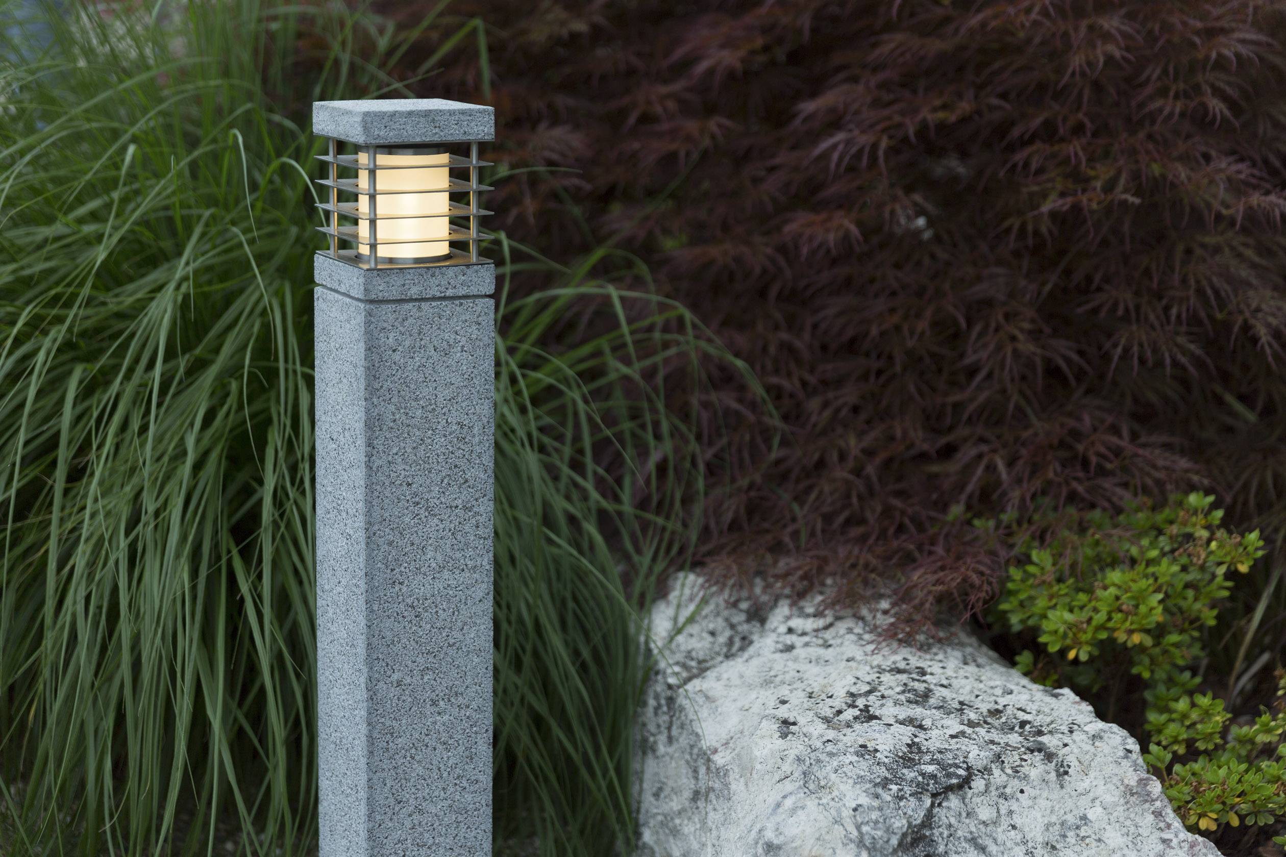 En modern golvlampa av sten lyser i en trädgård, omgiven av dekorativa växter och en stor sten.
