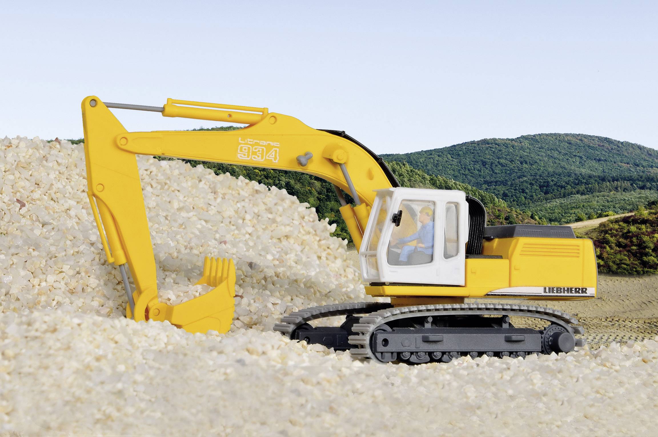 En gul grävmaskin i en grustäkt framför ett landskap med gröna kullar. Grävmaskinen lyfter material med sin skopa.