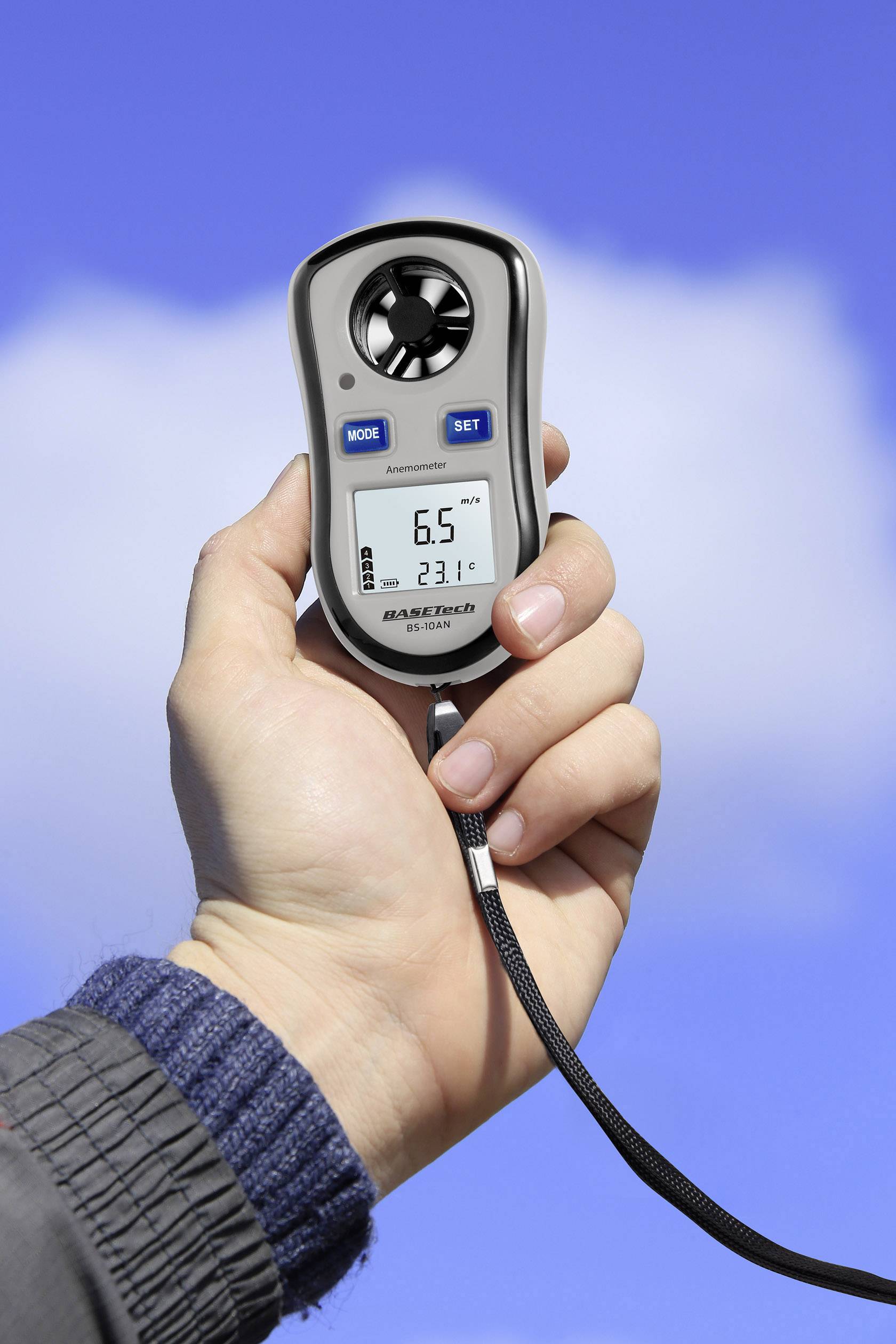 En hand håller ett anemometer i luften. Enheten visar en vindhastighet på 6,5 meter per sekund och en temperatur på 23,1 grader Celsius. Blå himmel i bakgrunden.