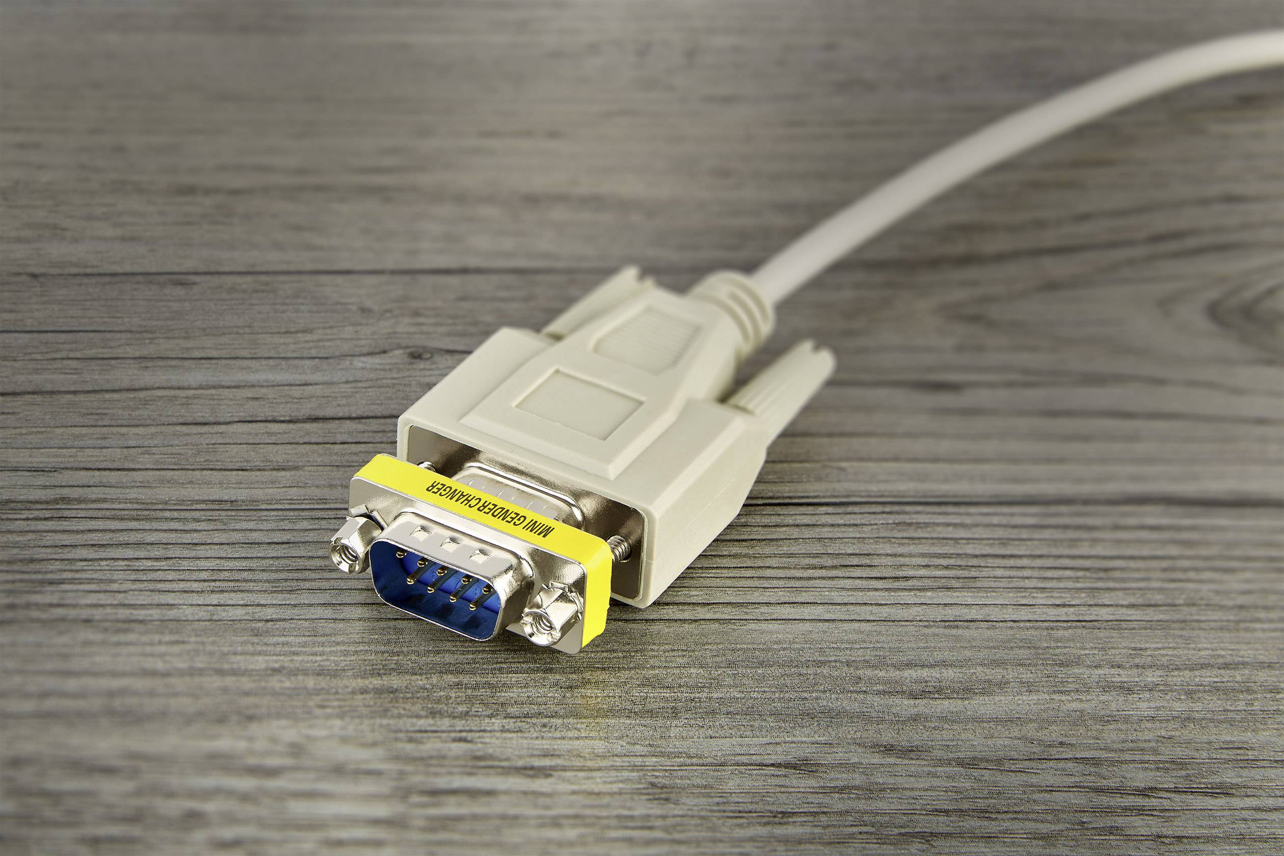 Ett grått VGA-kabel ligger på ett träbord. Det har blå stift och är lindad med en gul etikett som visar 'REMOVE BEFORE USE'.