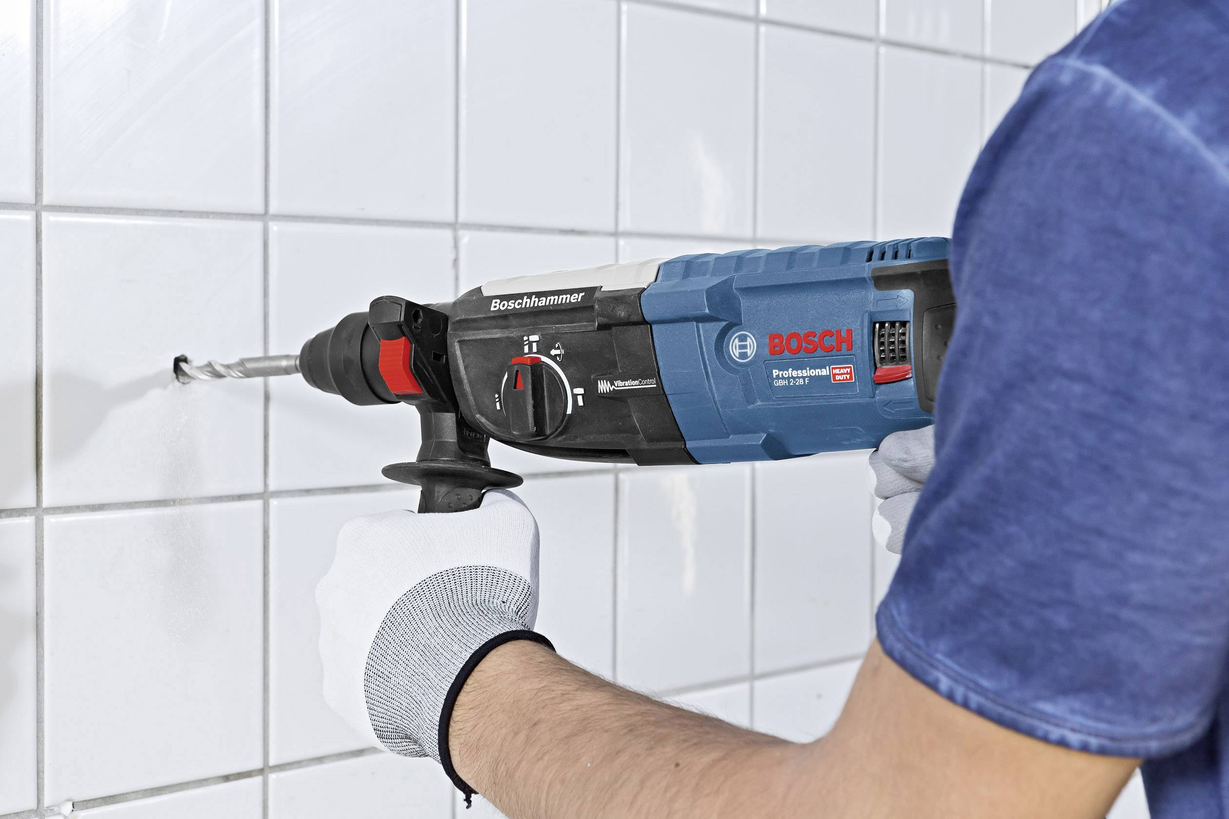 En person borrar ett hål i en vit kakelvägg med en blå Bosch-borr, och bär vita handskar.