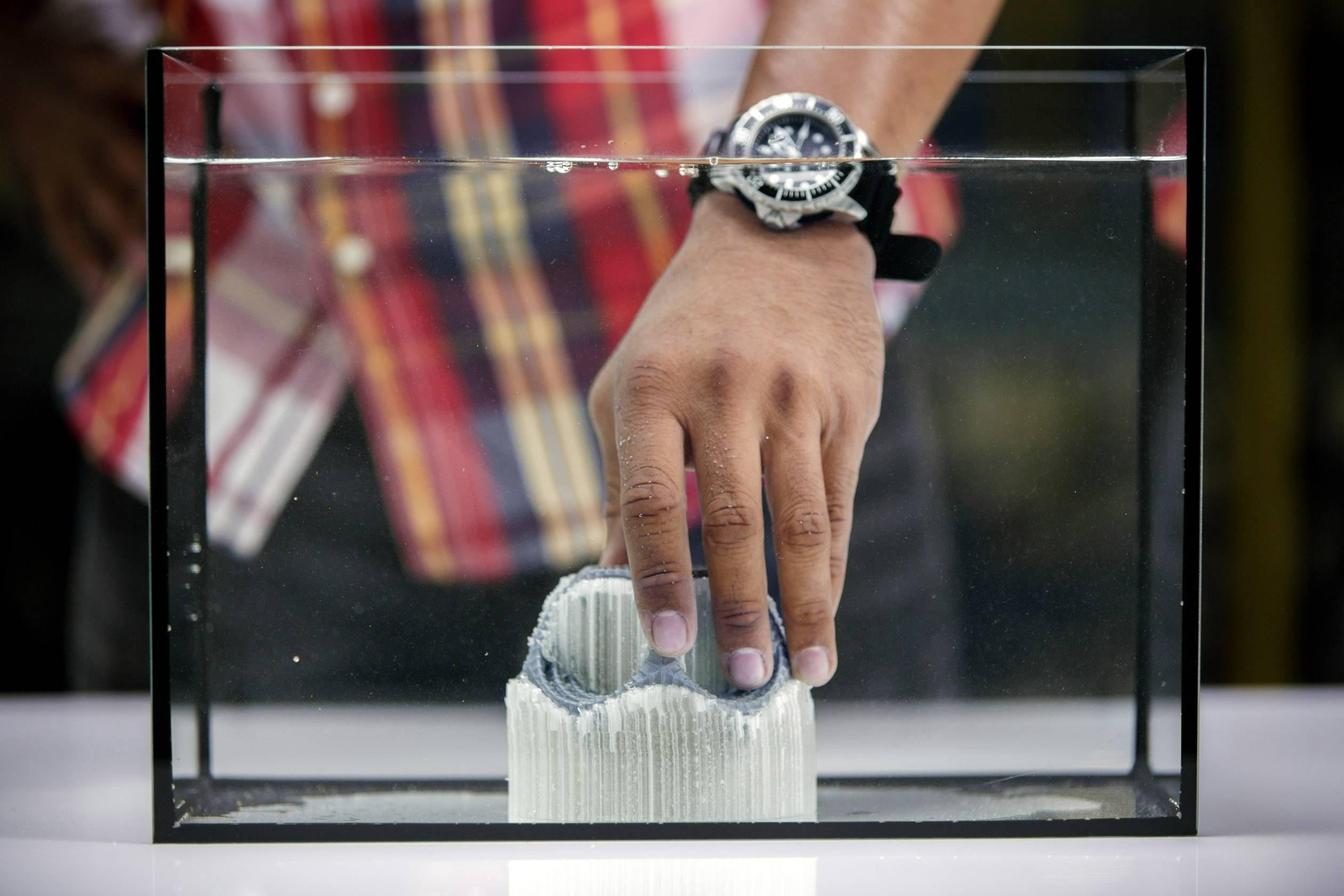 En hand med en klocka sänks ner i ett akvarium med en grå, porös struktur. Personen bär en rutig skjorta.