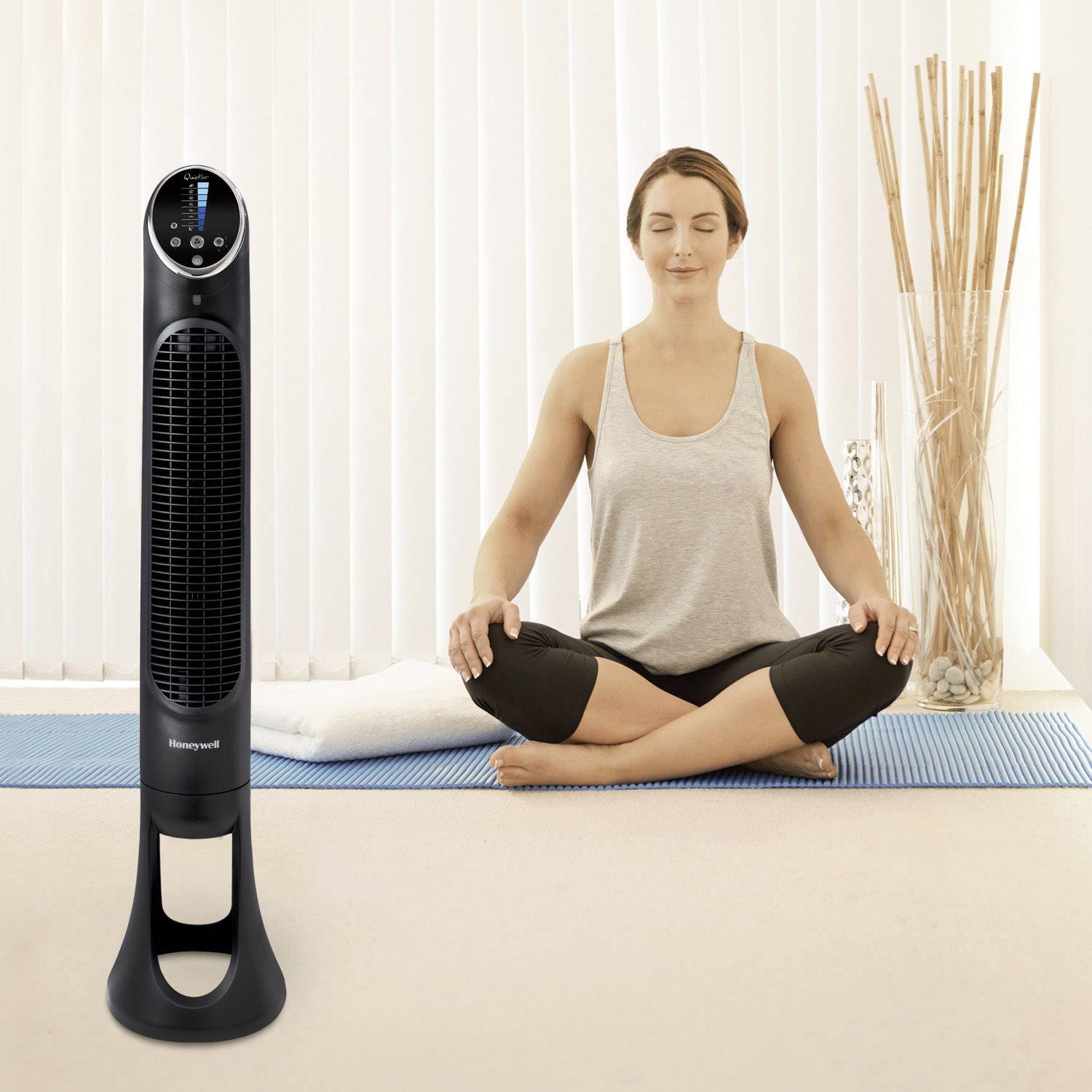 En kvinna sitter i taillös sits på en yogamatta, bredvid står en tornfläkt. Hon ser avslappnad ut.