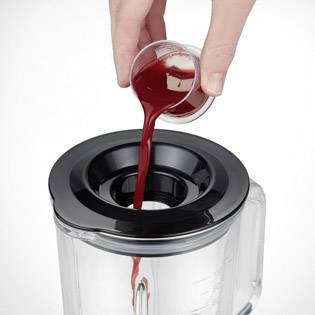 En hand häller röd vätska från en liten kanna i en öppen mixer.